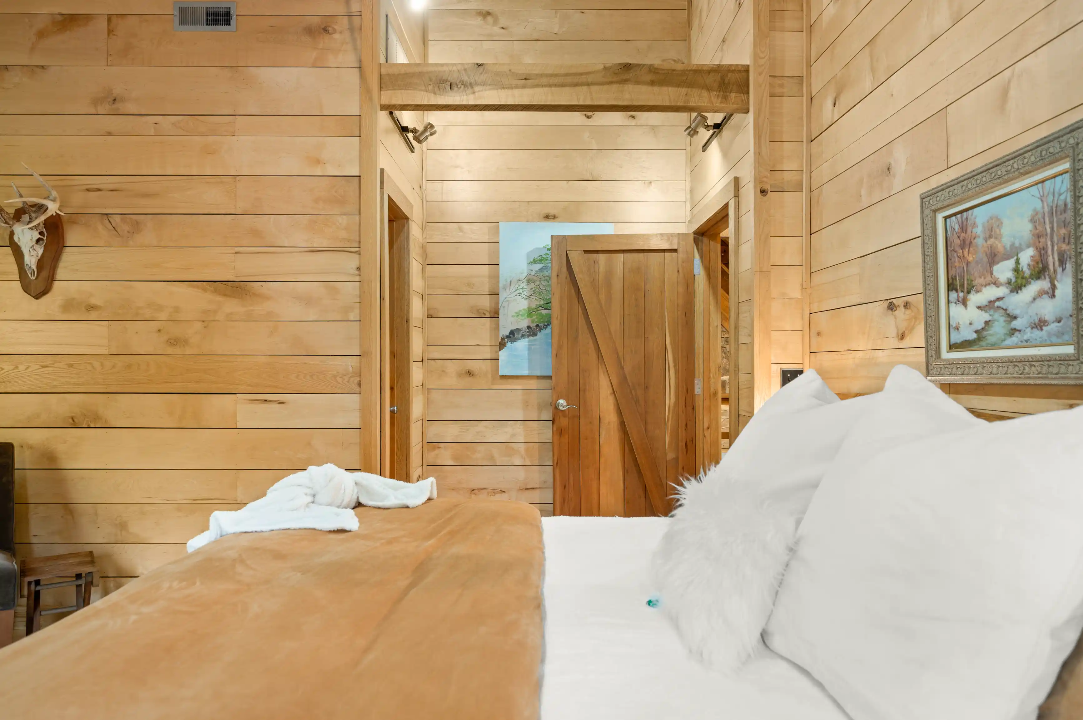 Cozy wooden cabin interior with a plush bed in the foreground, decorative framed artwork on the walls, and an open door leading to another room.