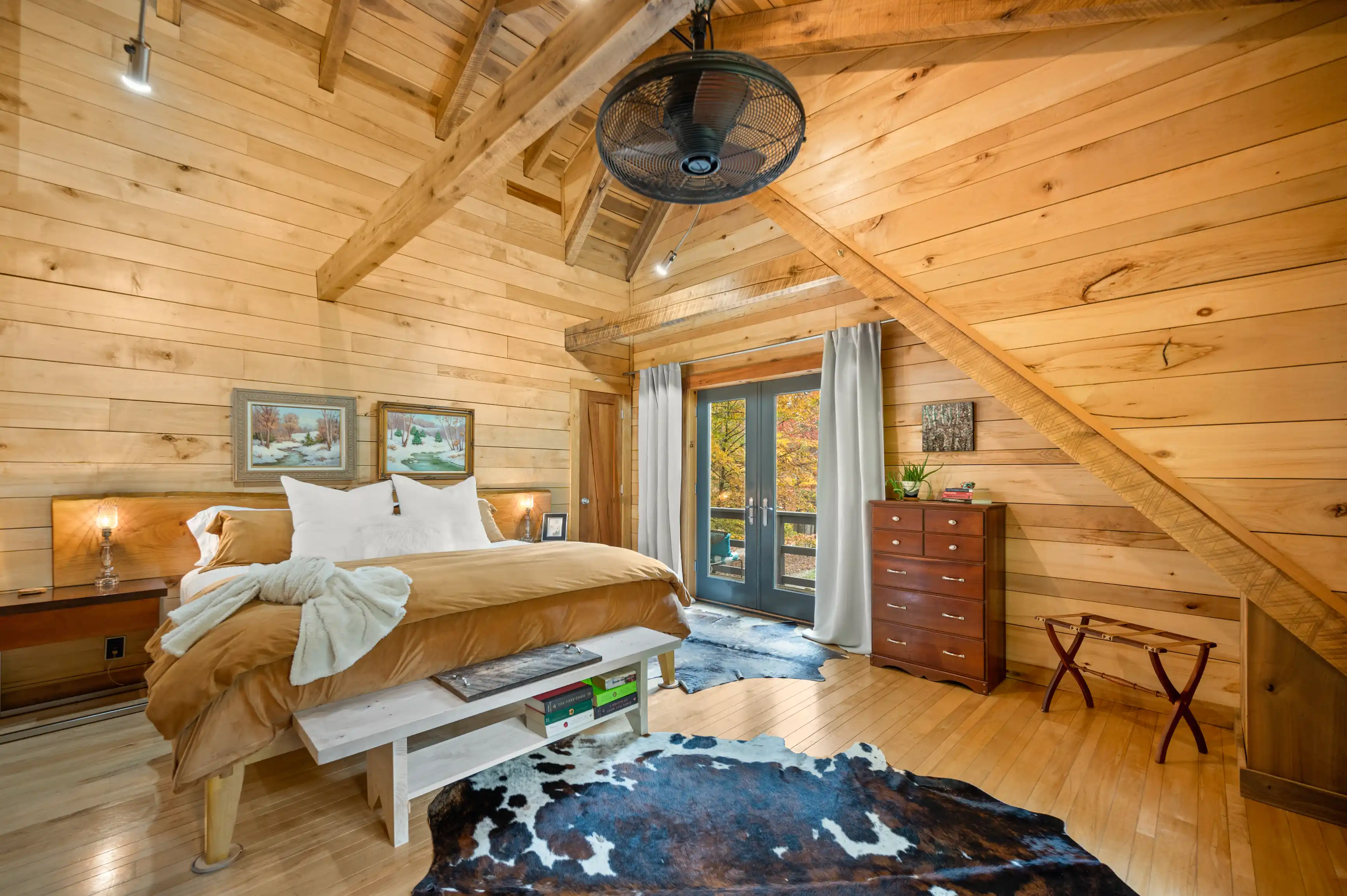 Cozy wooden cabin bedroom with a plush bed, cowhide rug, ceiling fan, and autumn view through the French doors.