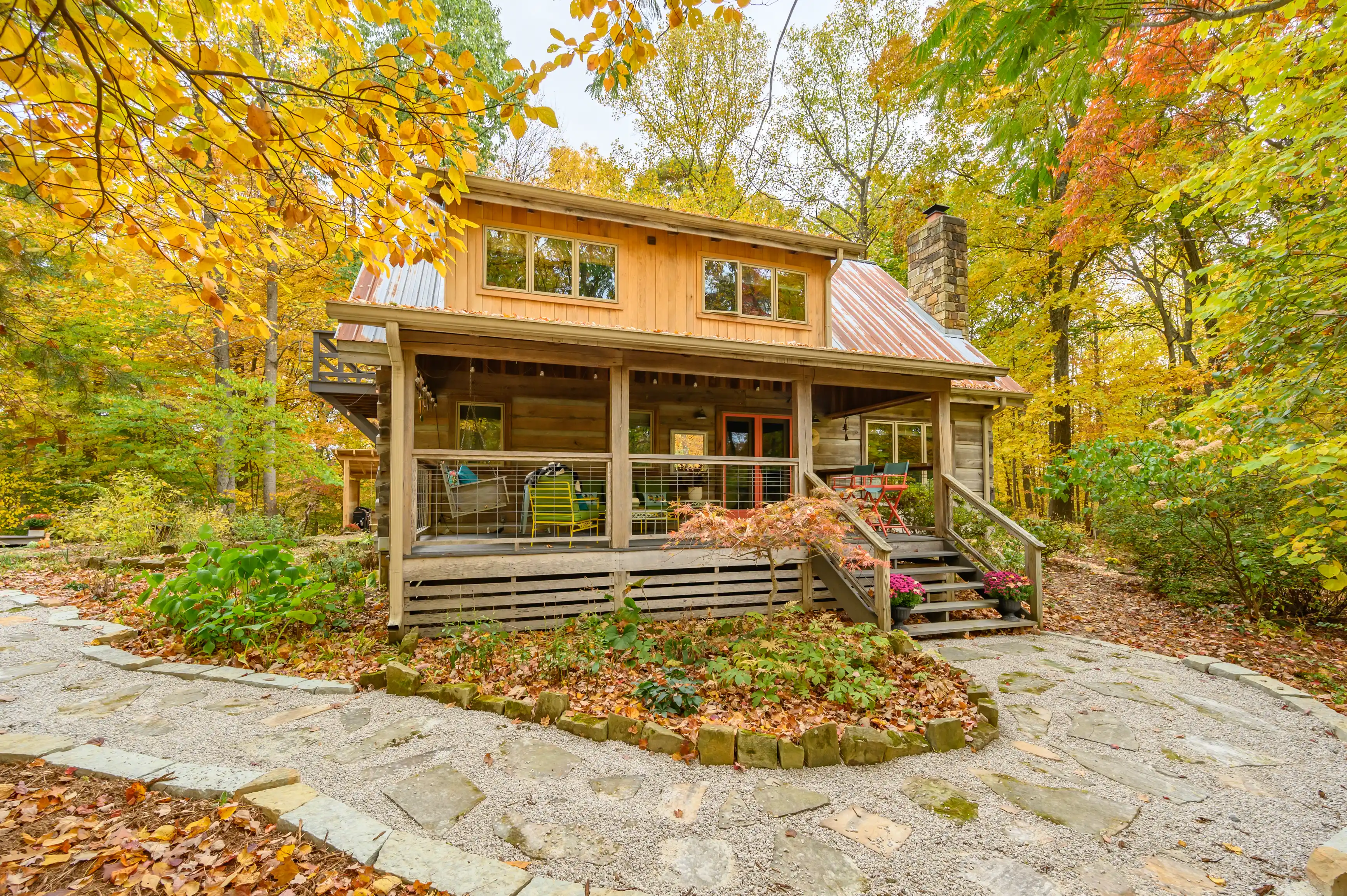 Quaint wooden cabin with a red tin roof surrounded by colorful fall foliage, featuring a front porch with a stone pathway.