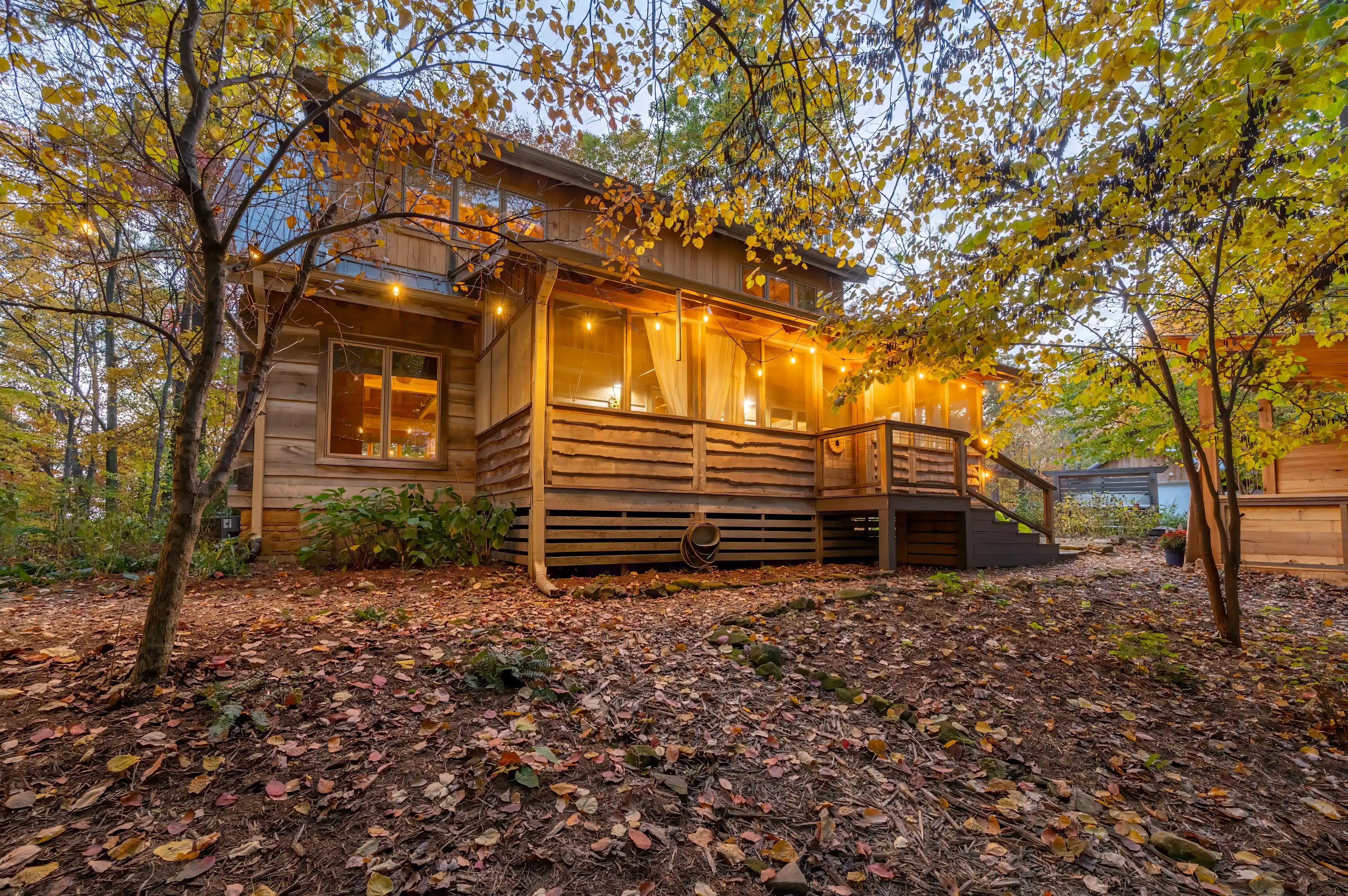 Cozy wooden cabin surrounded by autumn foliage with warm lights on at dusk.