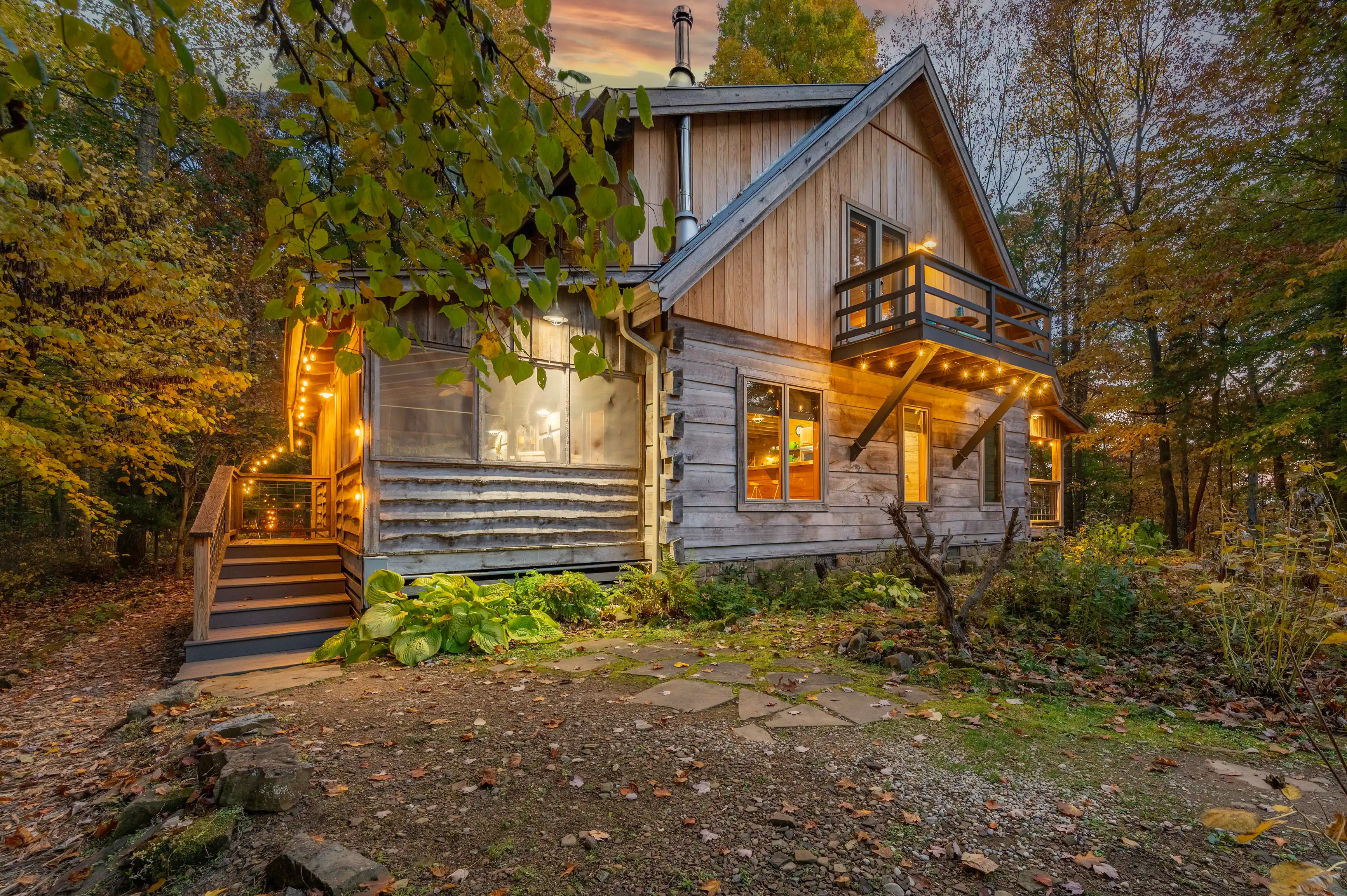 Rustic wooden cabin with illuminated porch lights, surrounded by autumn foliage at twilight.