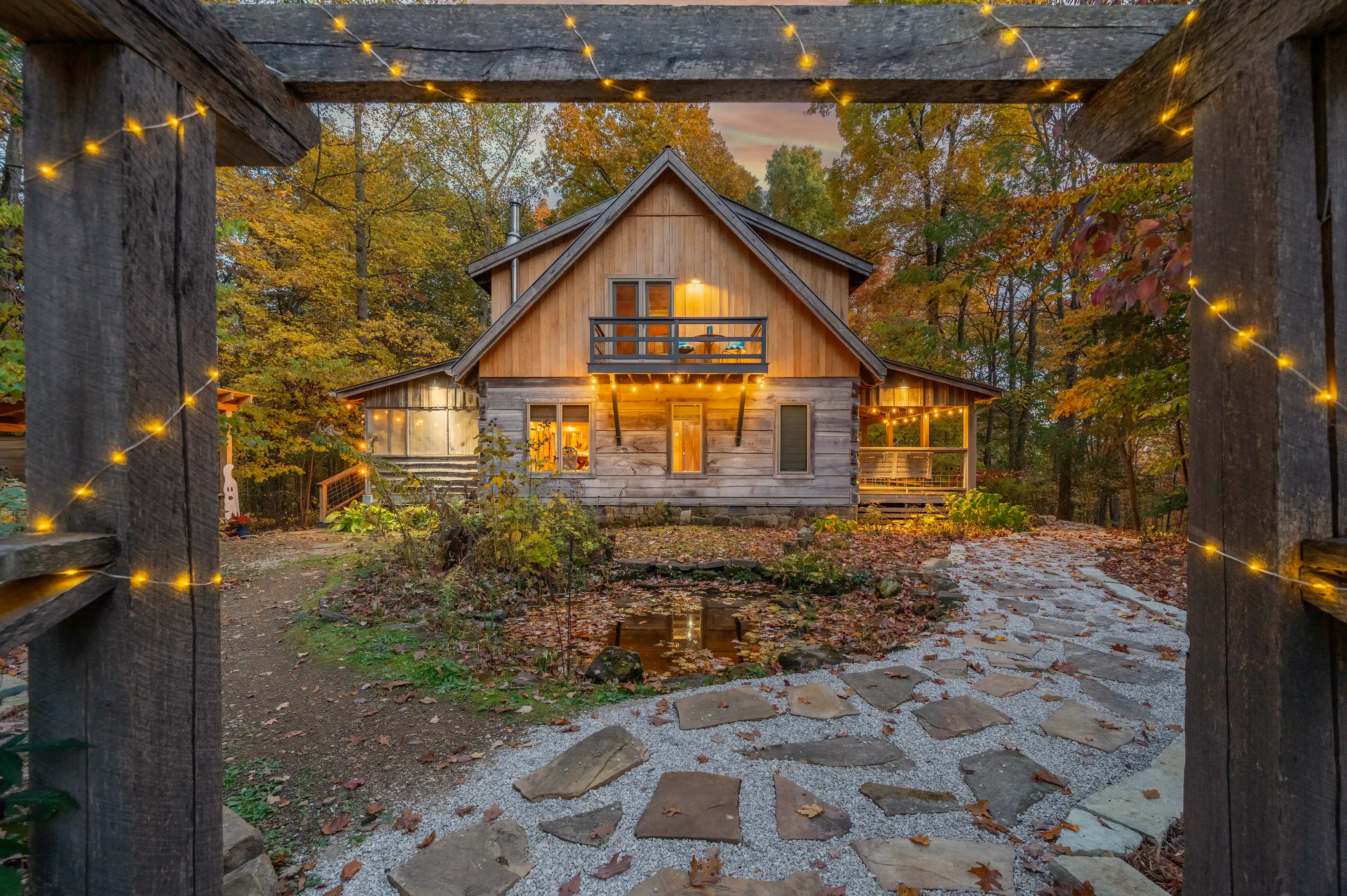 Cozy wooden cabin with lit interior and string lights at dusk, framed by a pergola, surrounded by autumn foliage.
