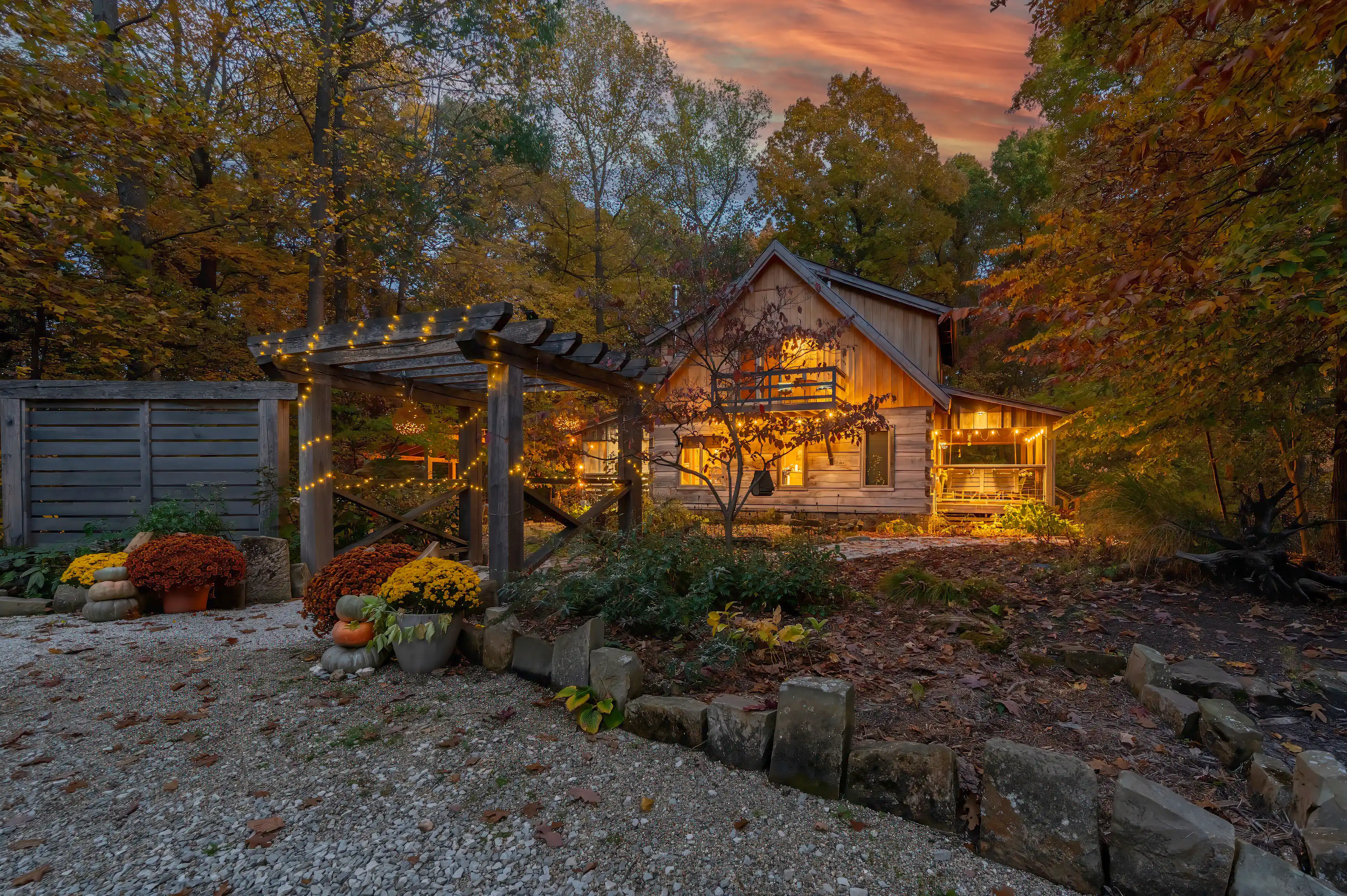 Cozy wooden cabin with warm lights at twilight surrounded by autumn foliage and decorated with potted mums and pumpkins.