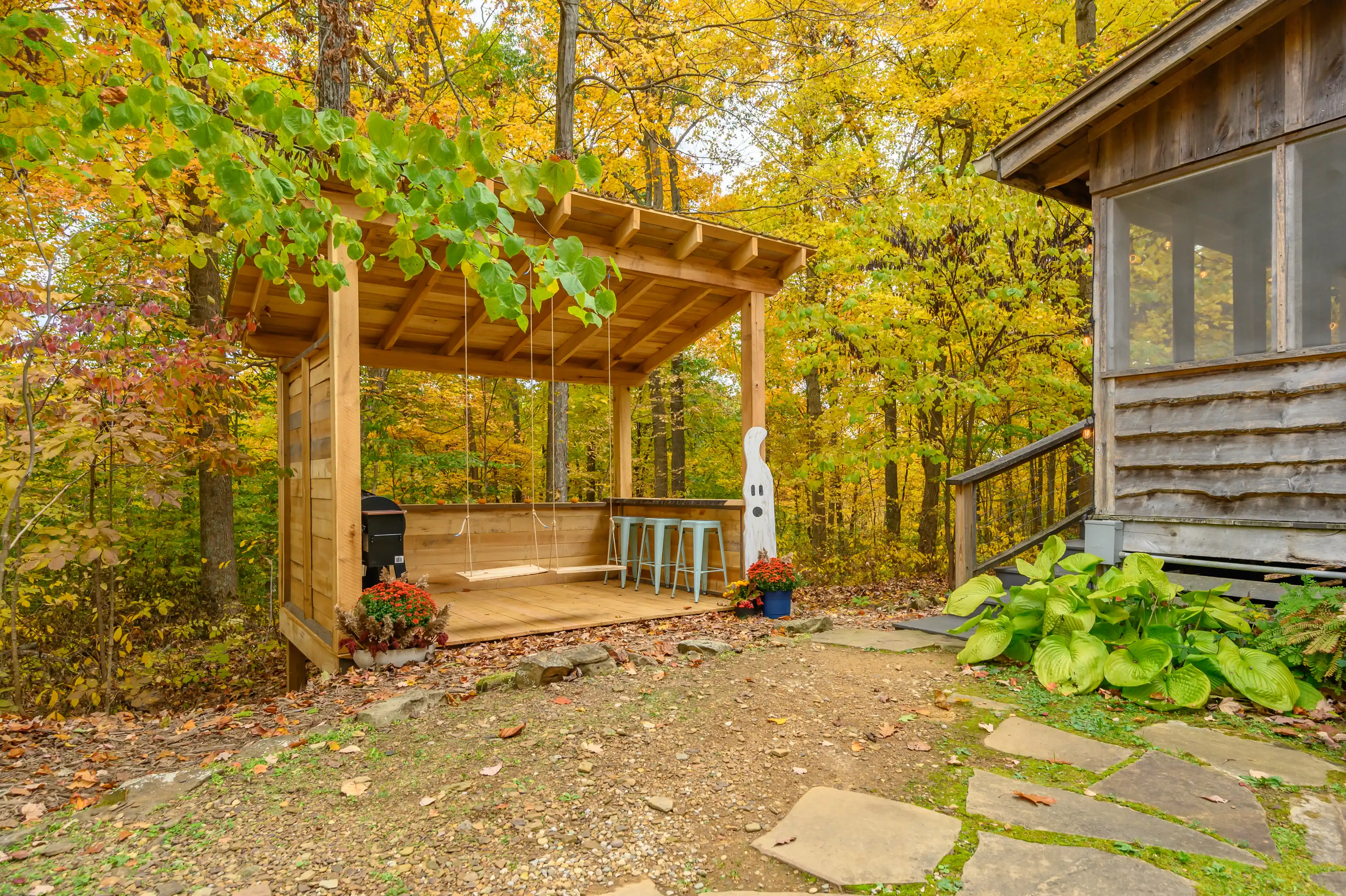 Wooden cabin with adjacent covered patio area in a forest with autumn foliage, featuring a swing, bar stools, and seasonal decorations.