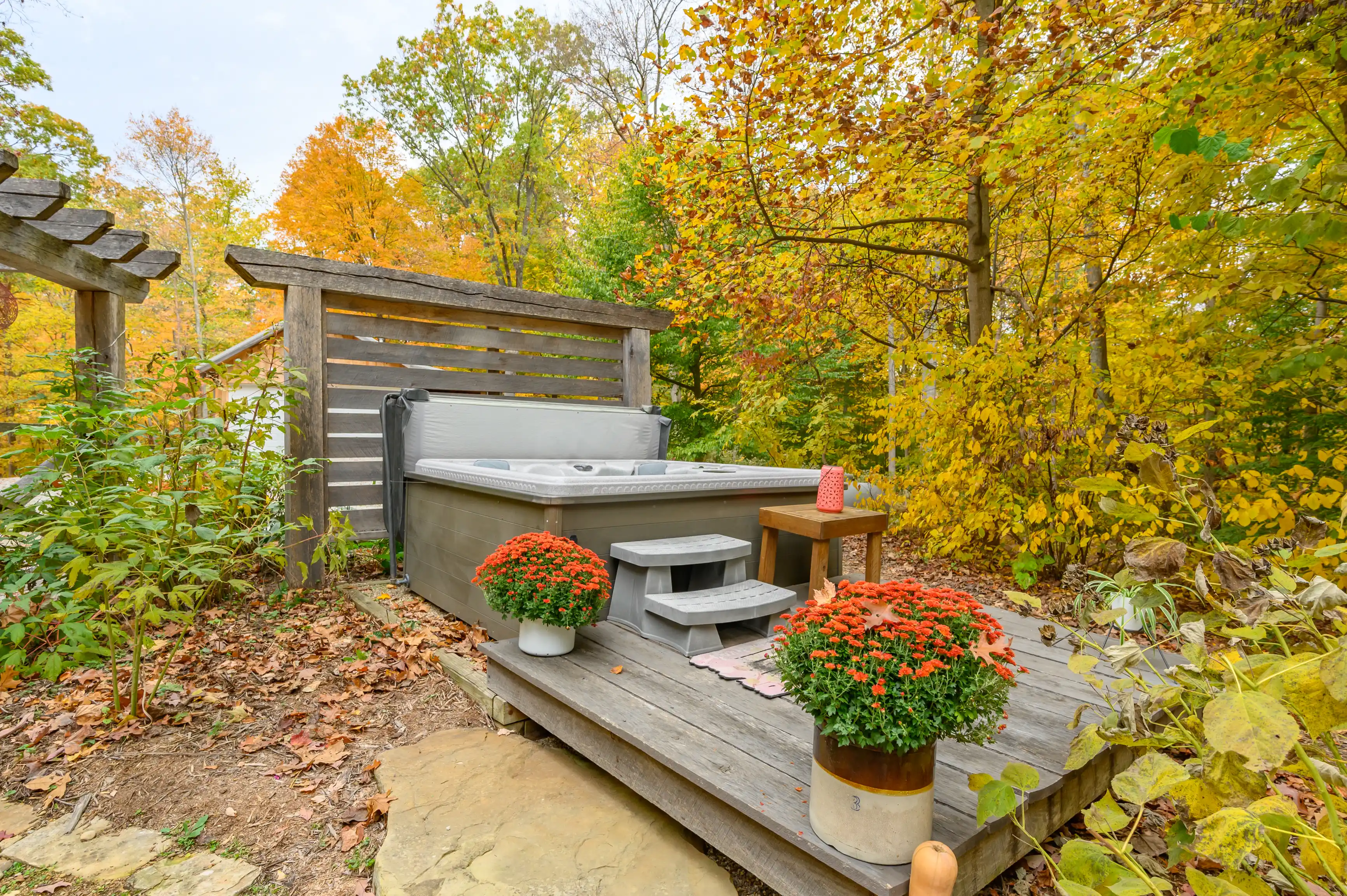 Outdoor hot tub on a wooden deck with steps, surrounded by autumn-colored trees and pots of blooming chrysanthemums.