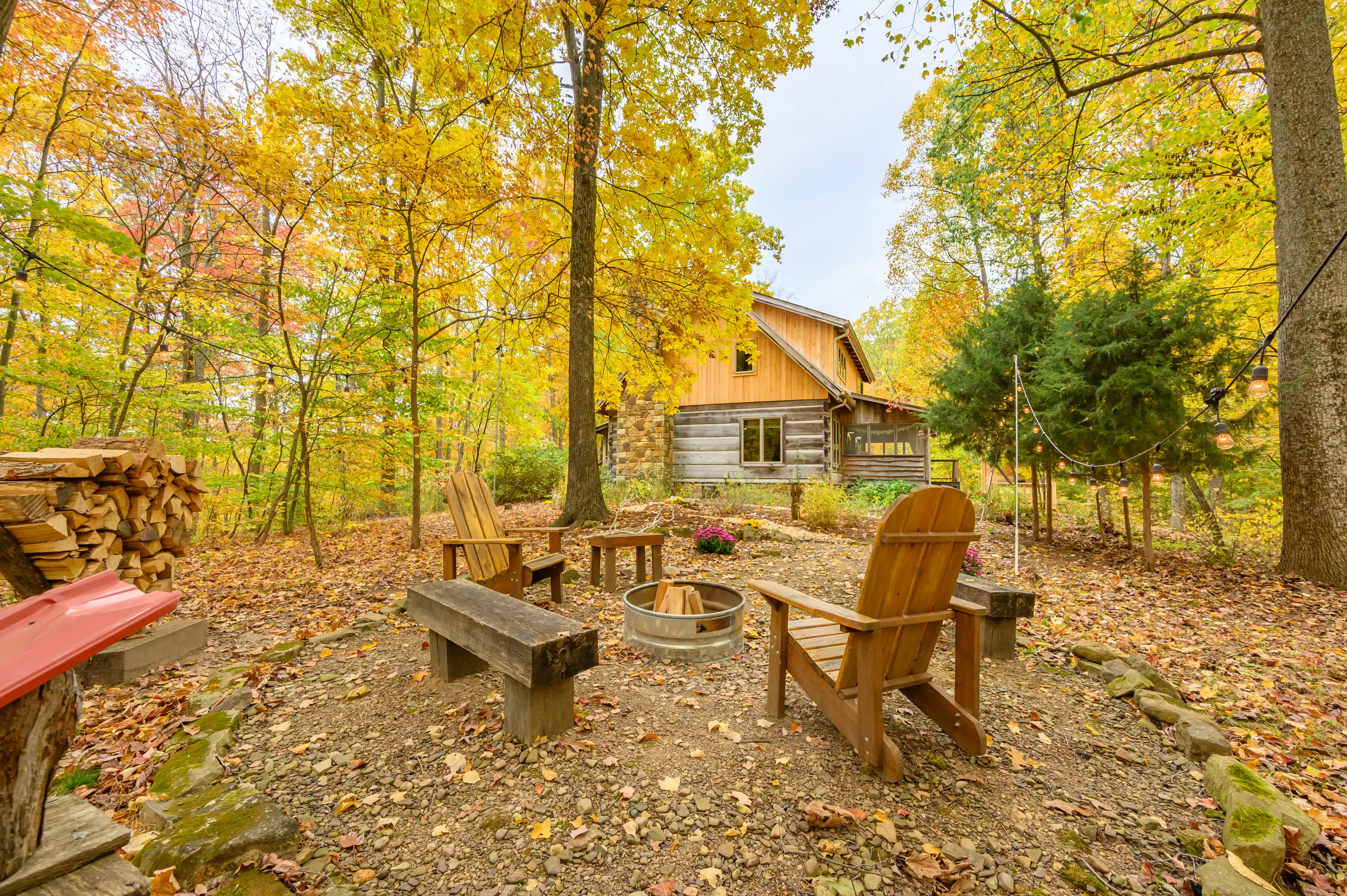 Rustic cabin surrounded by autumn-colored trees, featuring an outdoor seating area with Adirondack chairs and a fire pit.