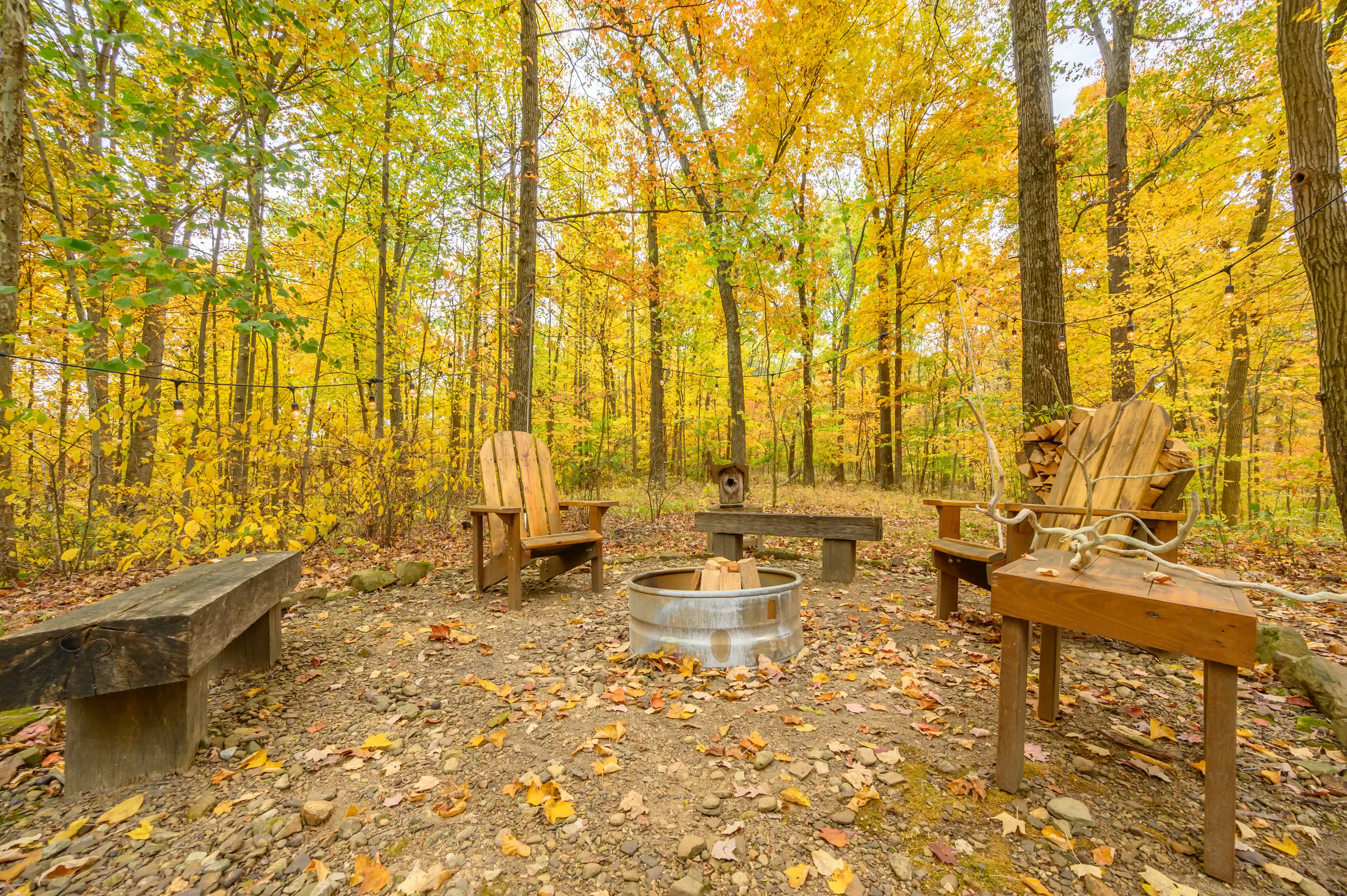 A cozy autumnal outdoor setting with Adirondack chairs around a fire pit in a forest with fall foliage.