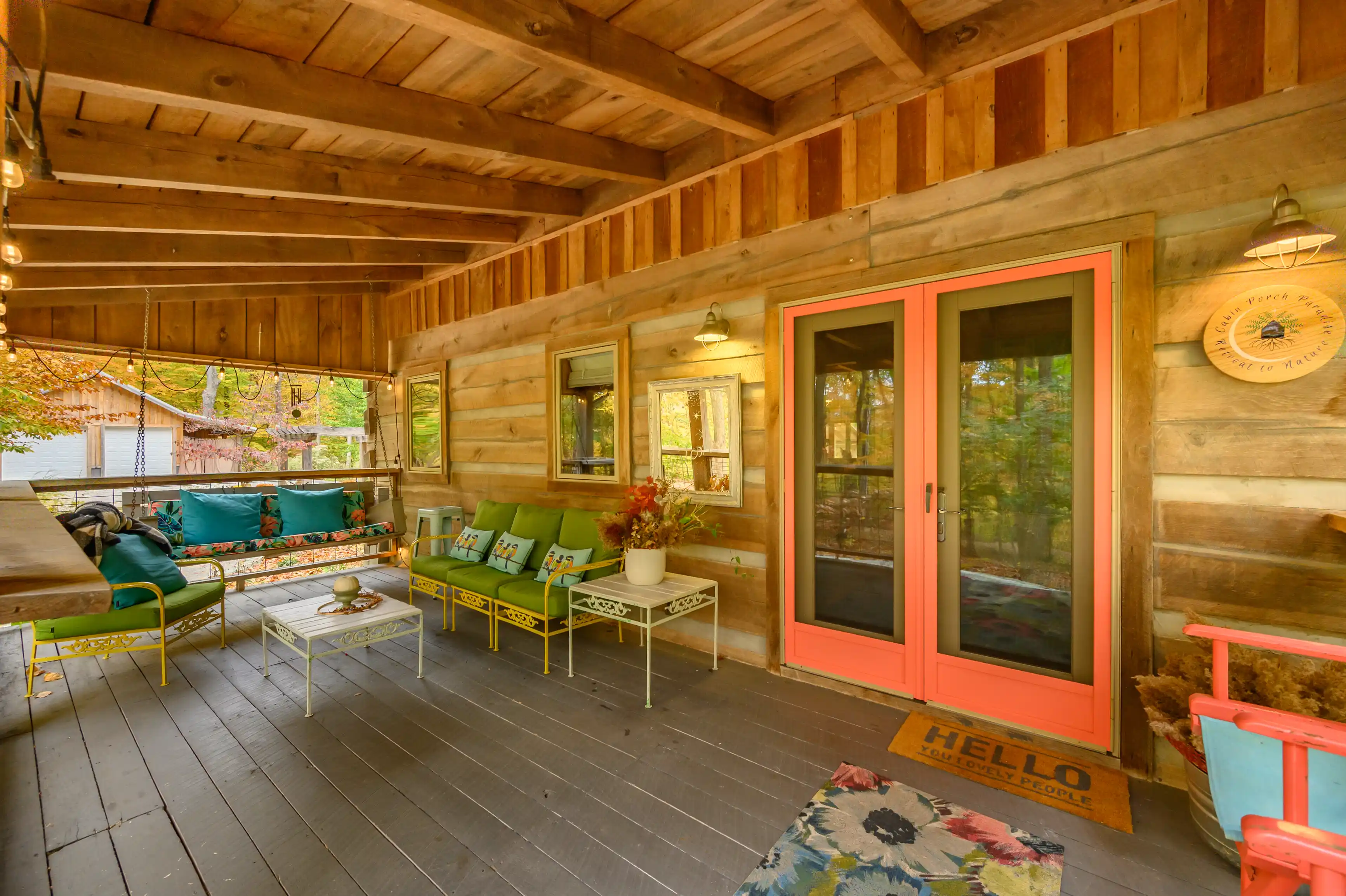Cozy covered porch area of a log cabin with colorful furniture, a "HELLO" doormat, and warm lighting.