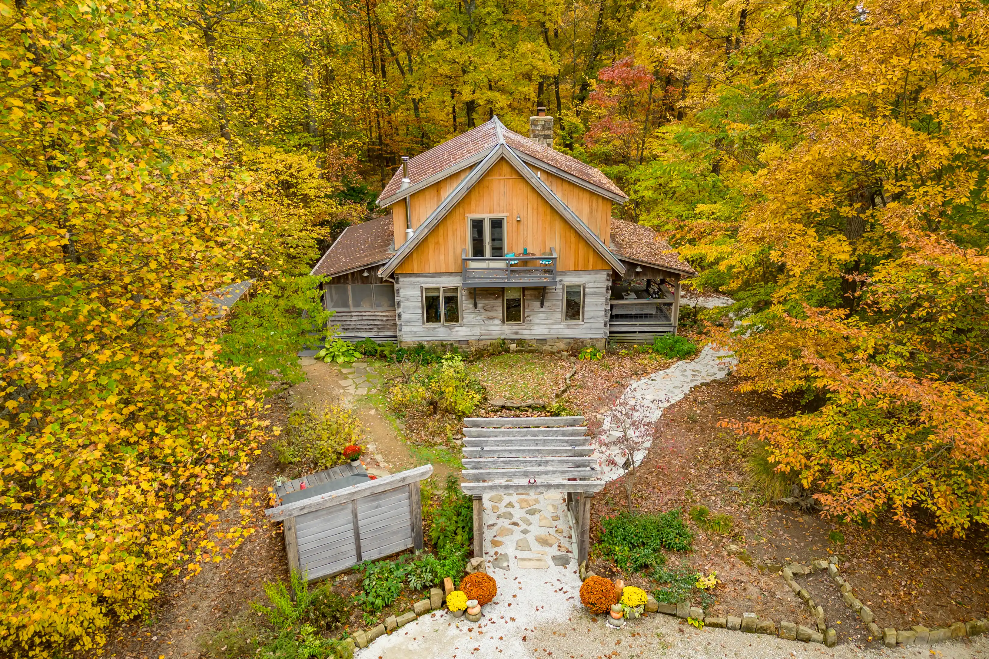 Aerial view of a quaint wooden cabin surrounded by lush autumn foliage with a path leading to a rustic entryway adorned with seasonal decorations.