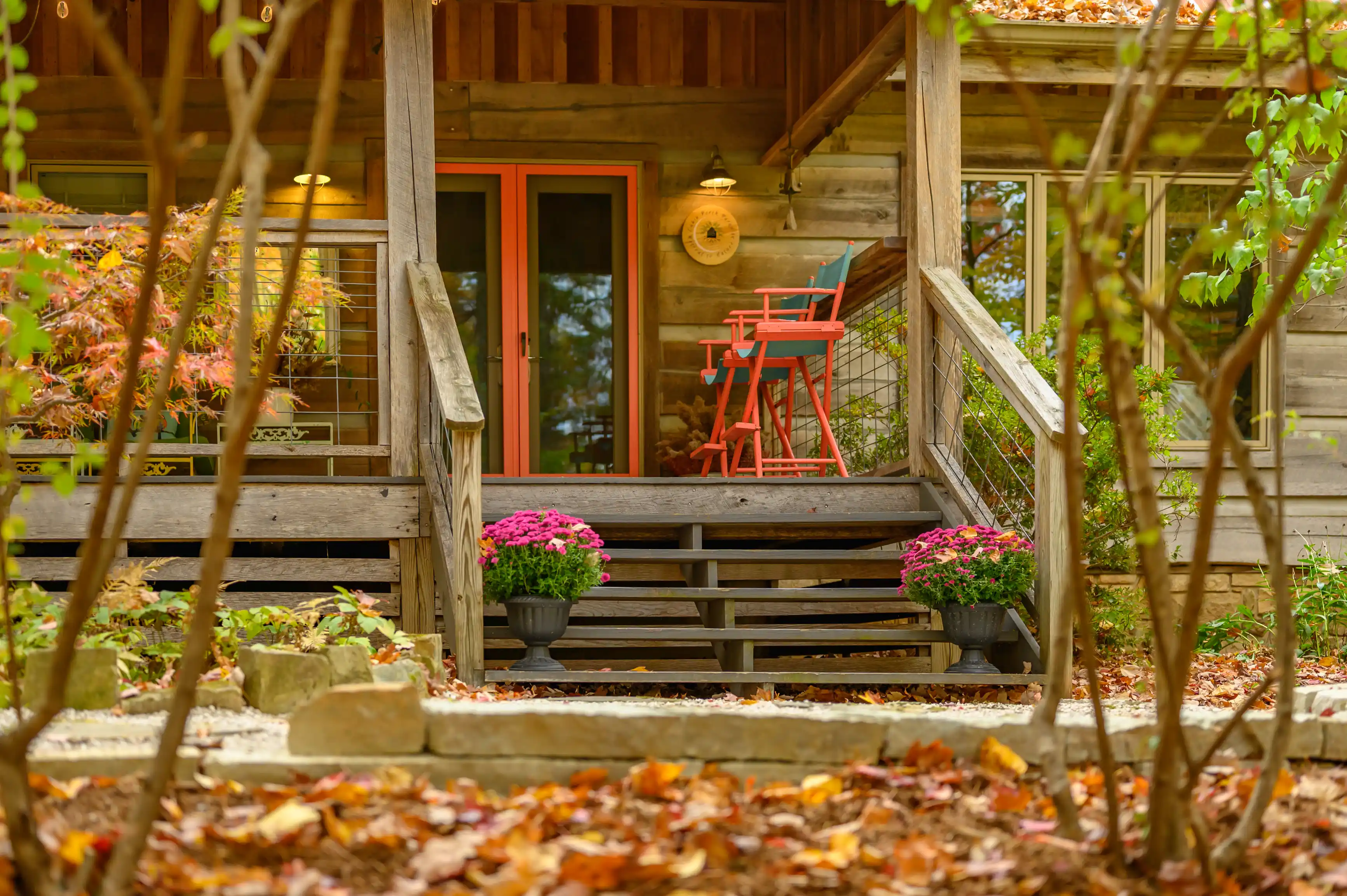 Cozy wooden cabin porch with orange double doors, red high chair, pink potted flowers, and fallen autumn leaves.