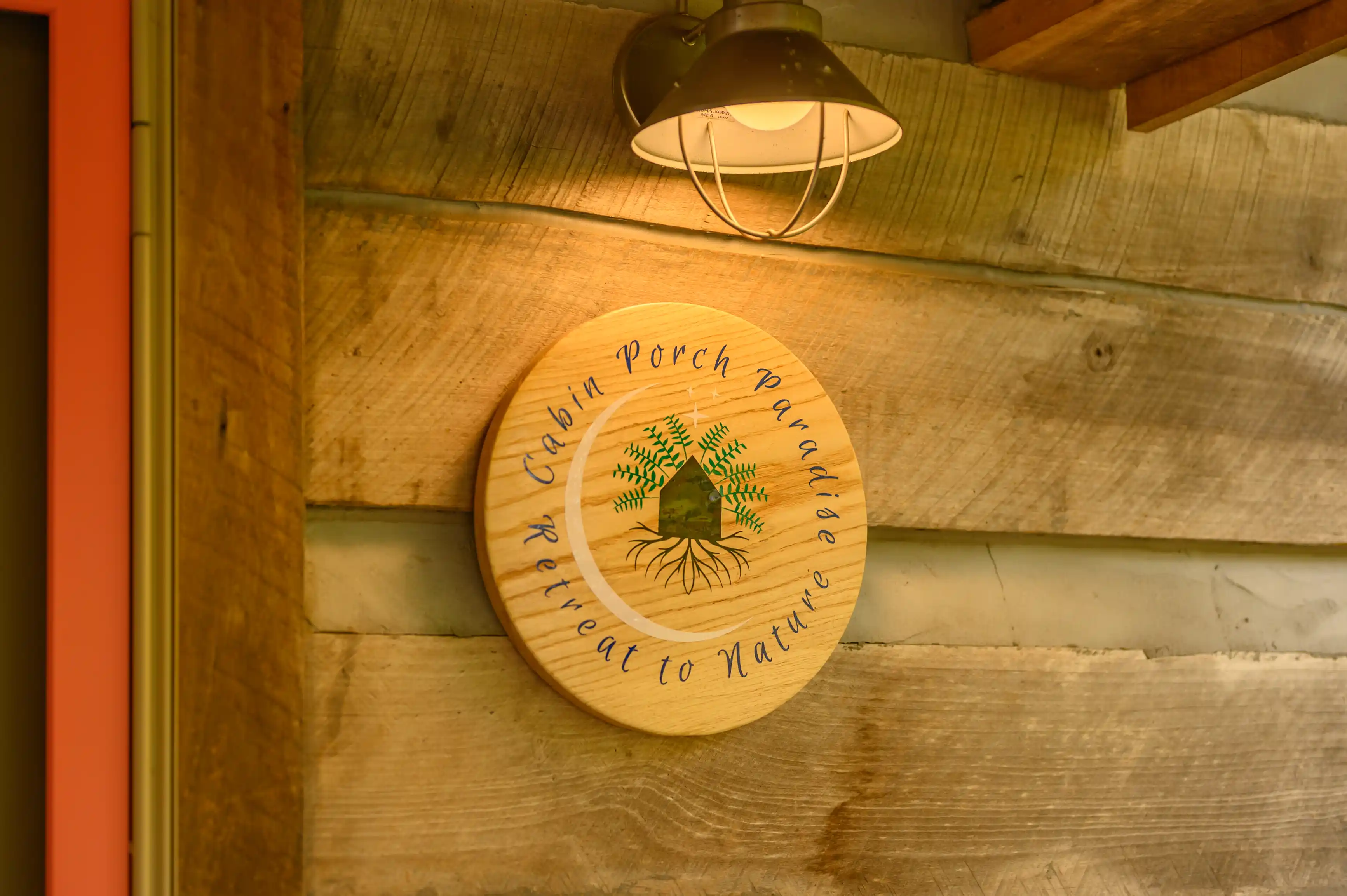 Round wooden sign with inscribed text "Cabin Porch Paradise, Retreat to Nature" on a rustic wall, illuminated by a vintage hanging lamp.