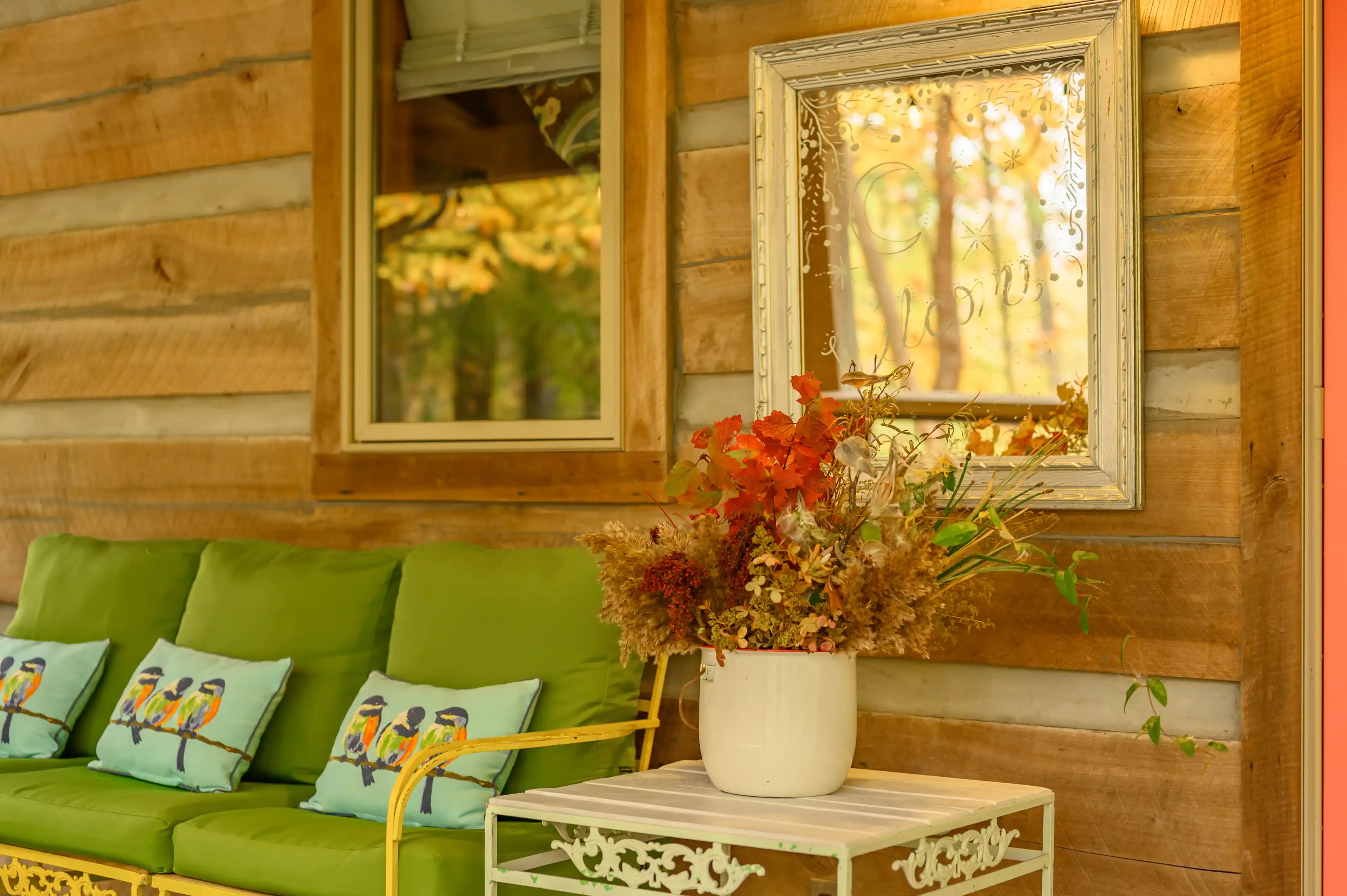 Cozy porch corner with green cushions on a bench, vibrant autumn flower arrangement in a white vase, and a reflective ornate mirror on a wooden wall.