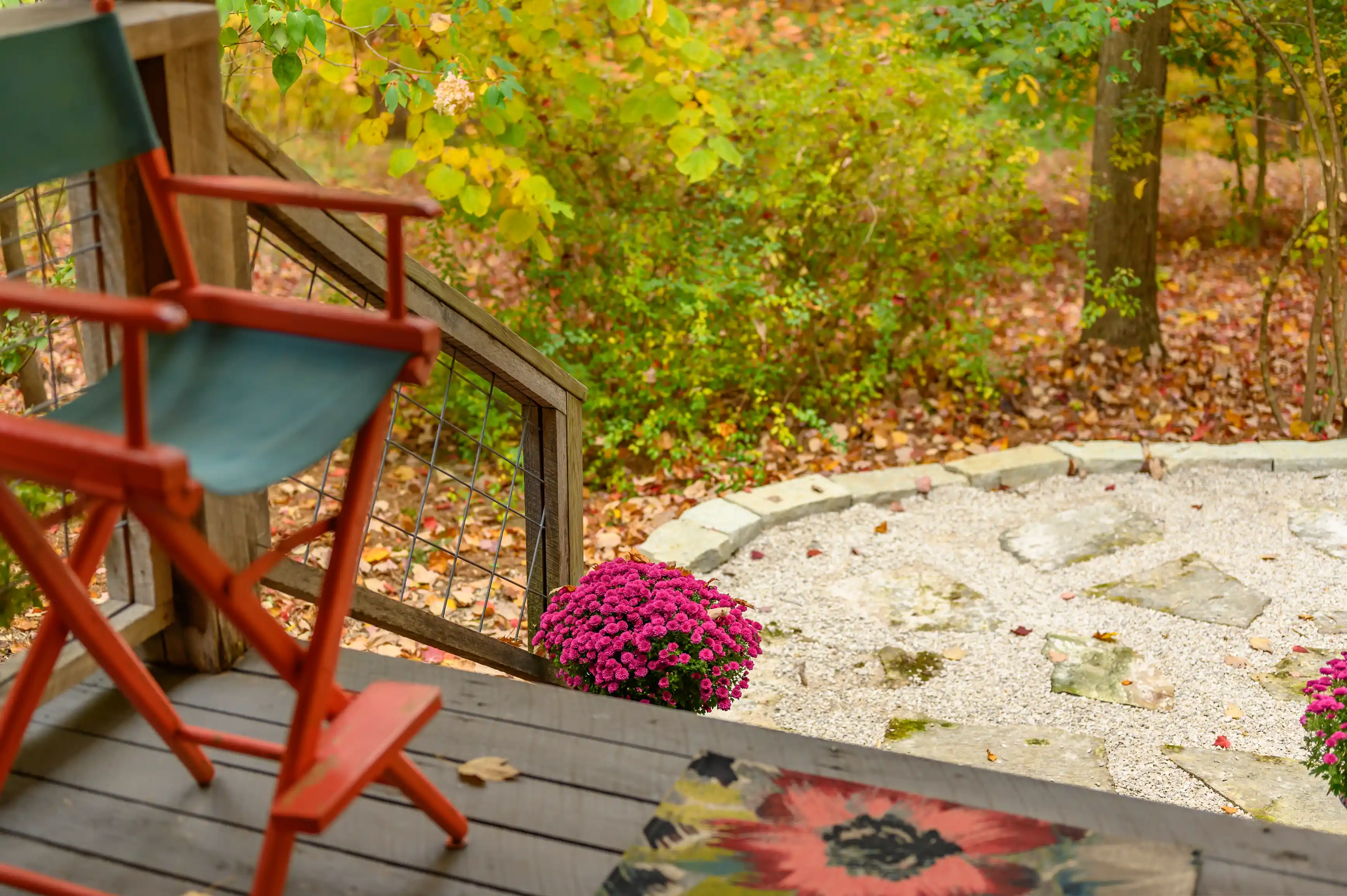 A cozy autumn porch with a red rocking chair overlooking a garden with blooming purple chrysanthemums and a stone pathway amidst fallen leaves.
