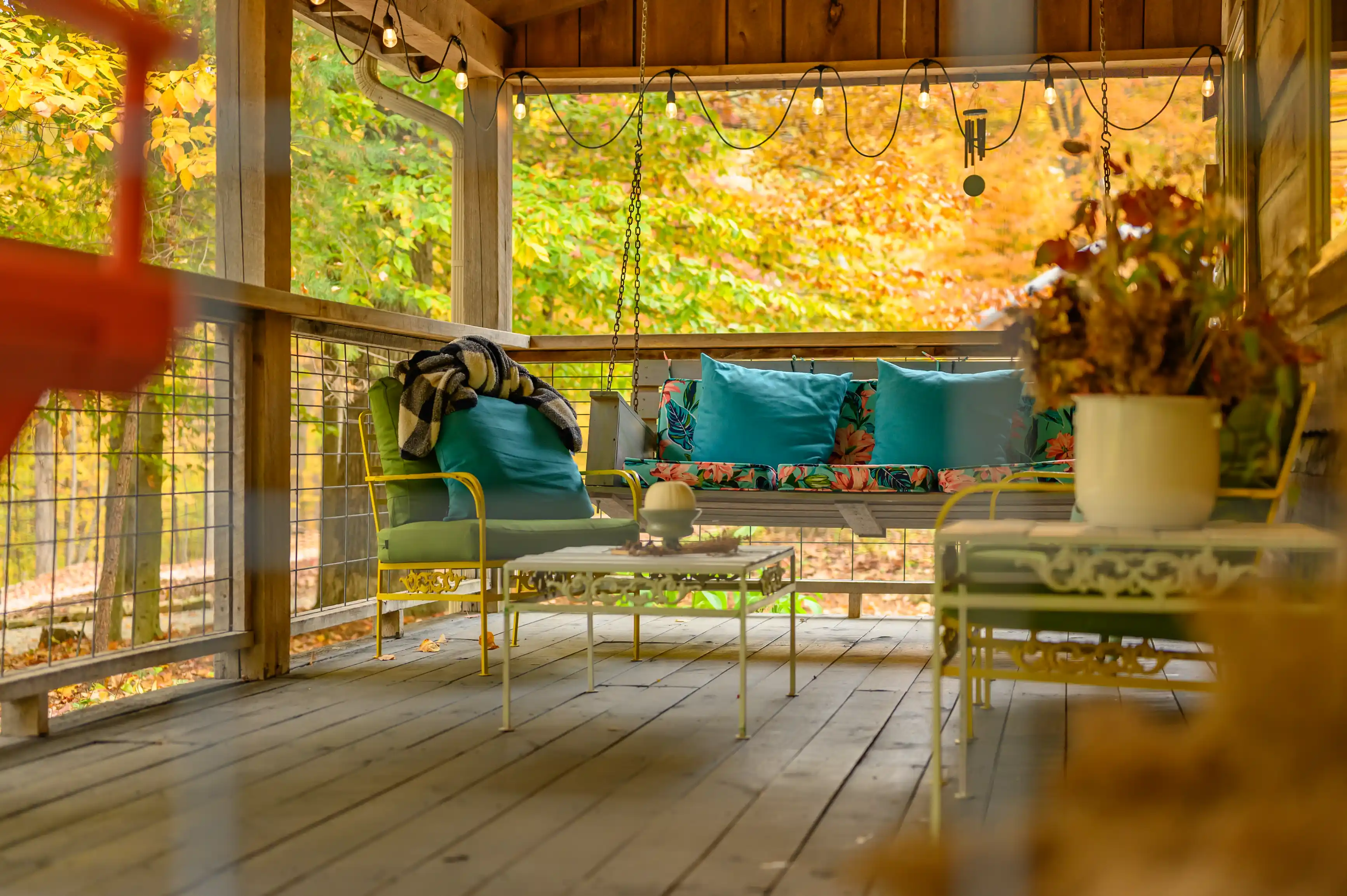 Cozy, covered porch with a swinging bench adorned with blue cushions and a plaid blanket, surrounded by autumnal trees, featuring string lights and a tabletop with a potted plant.