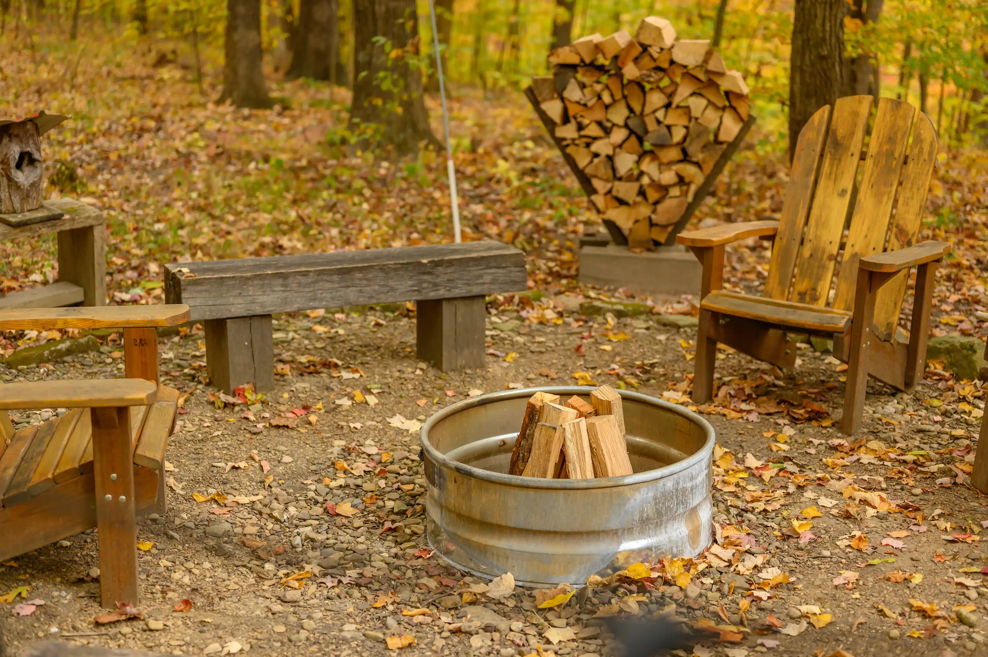 Outdoor fire pit with chopped firewood, surrounded by wooden benches and chairs, with autumn leaves scattered on the ground.