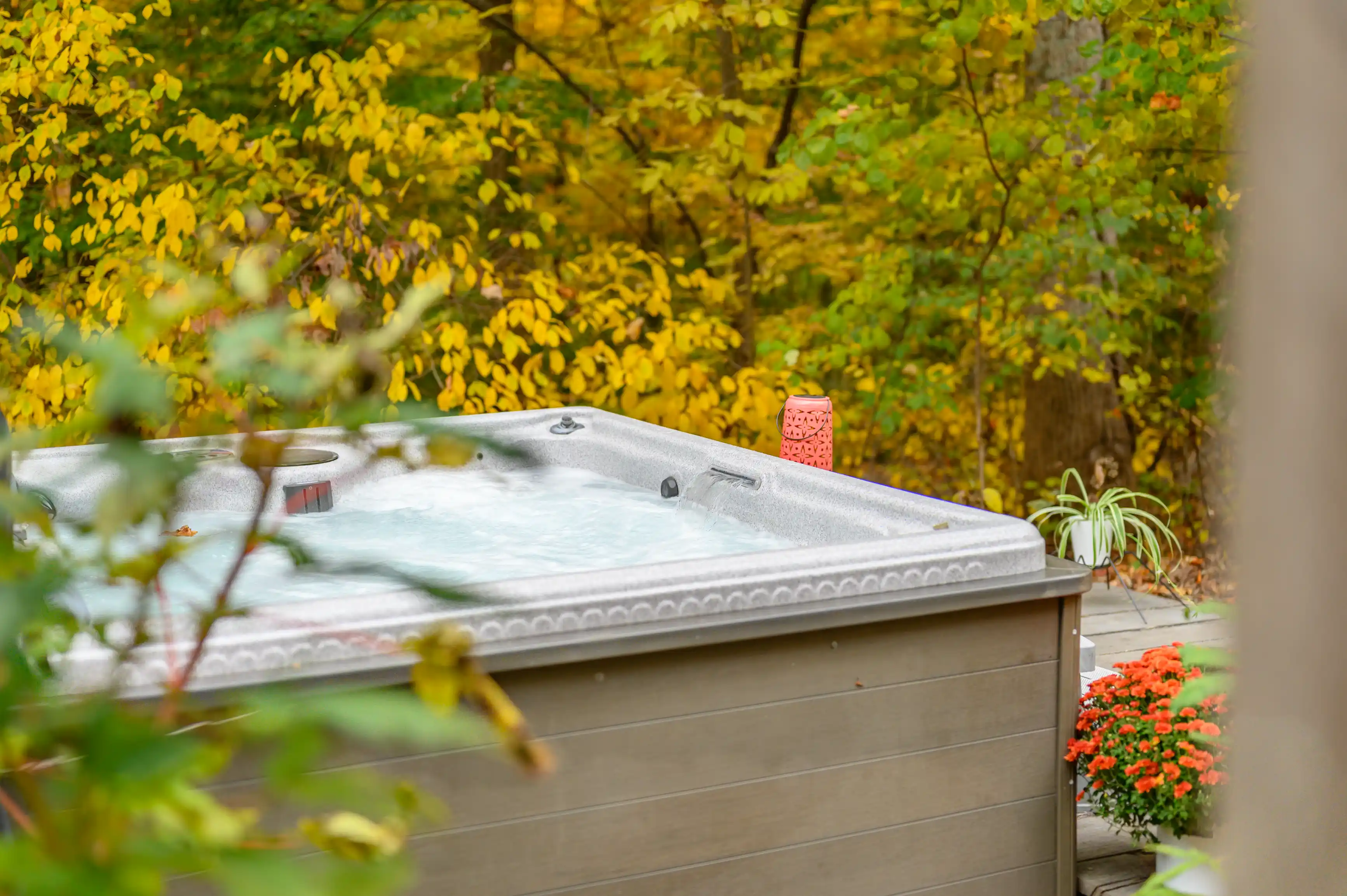 Outdoor hot tub with a cover on a wooden deck surrounded by autumn foliage and blooming flowers.