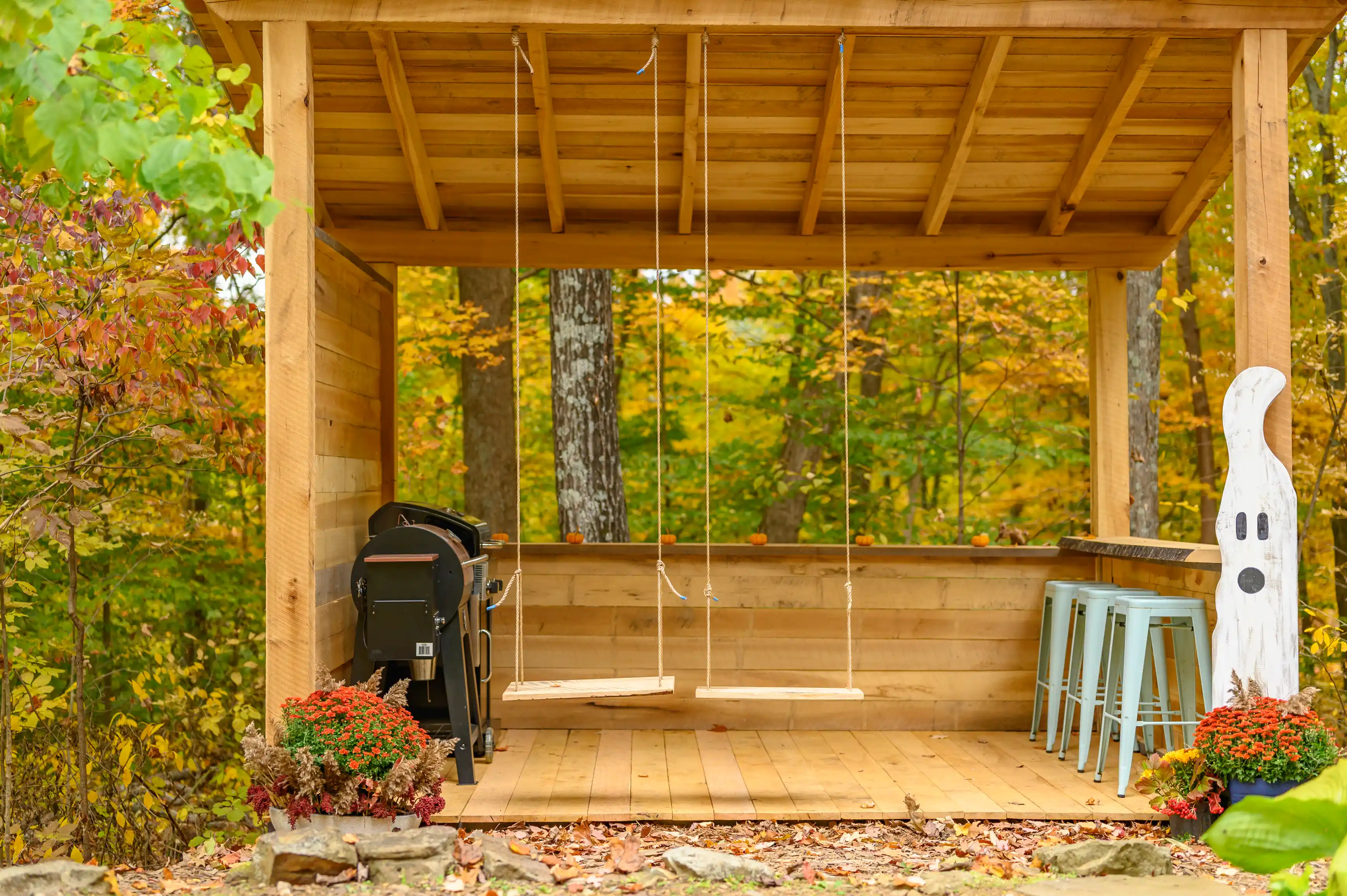 Wooden porch with a swing, barbecue grill, and Halloween decorations, surrounded by autumn foliage.