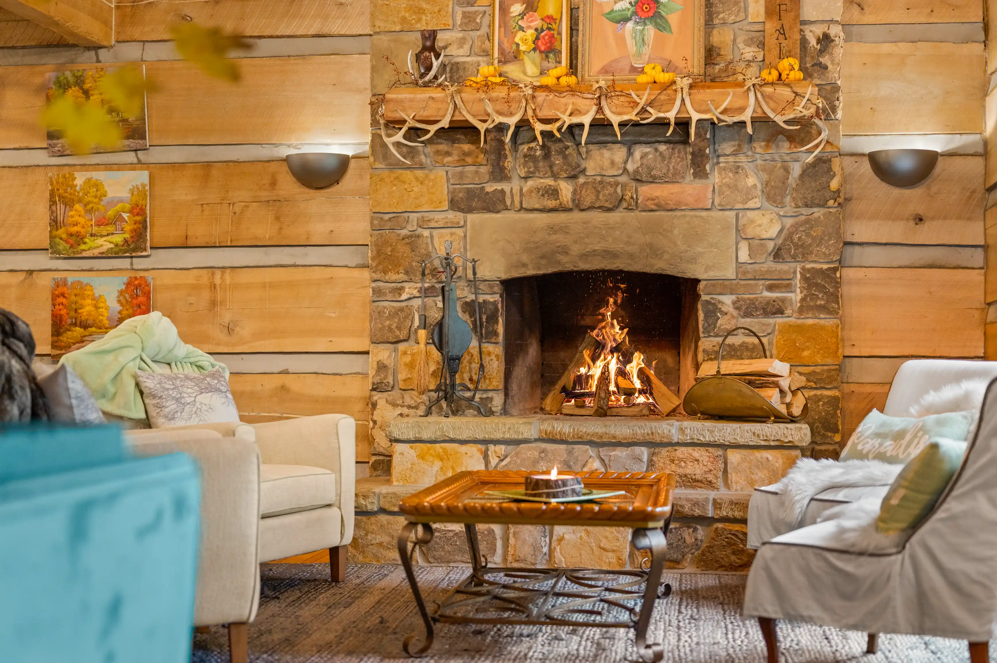 Cozy cabin interior with a lit fireplace, stone surround, wooden walls, framed autumn landscape paintings, decorative antlers, and comfortable seating area with a small wooden table.