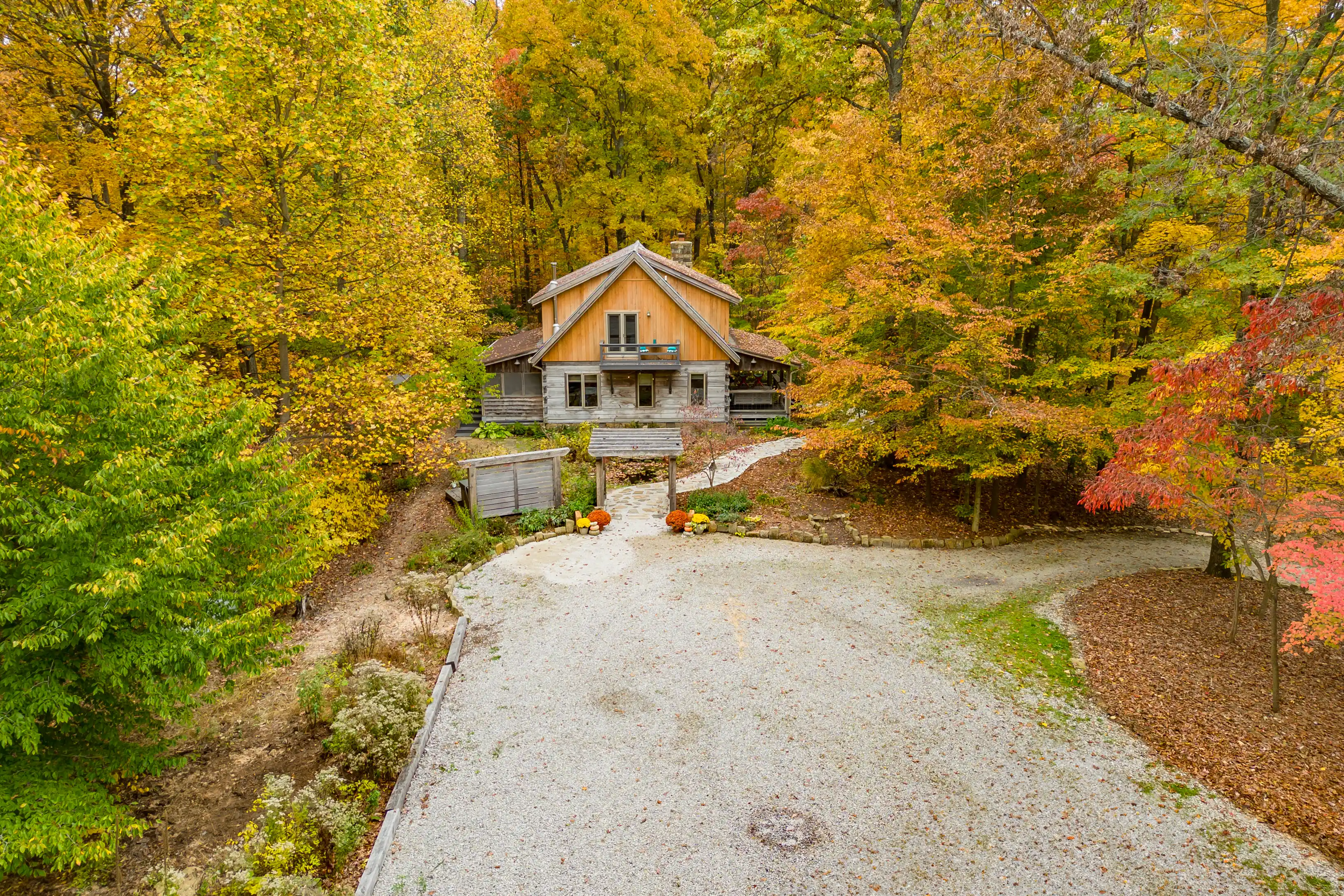 Cozy wooden cabin surrounded by a colorful autumn forest with a gravel driveway and pumpkins near the entrance.