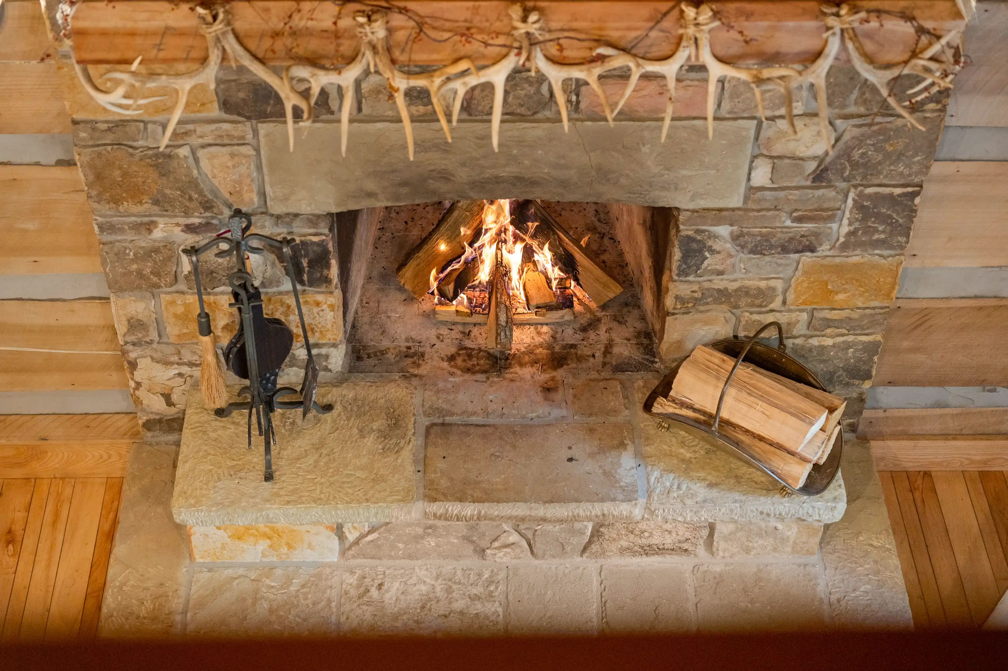 Cozy fireplace with a burning fire, flanked by fireplace tools on the left and a stack of logs on the right, under a mantel adorned with antlers.