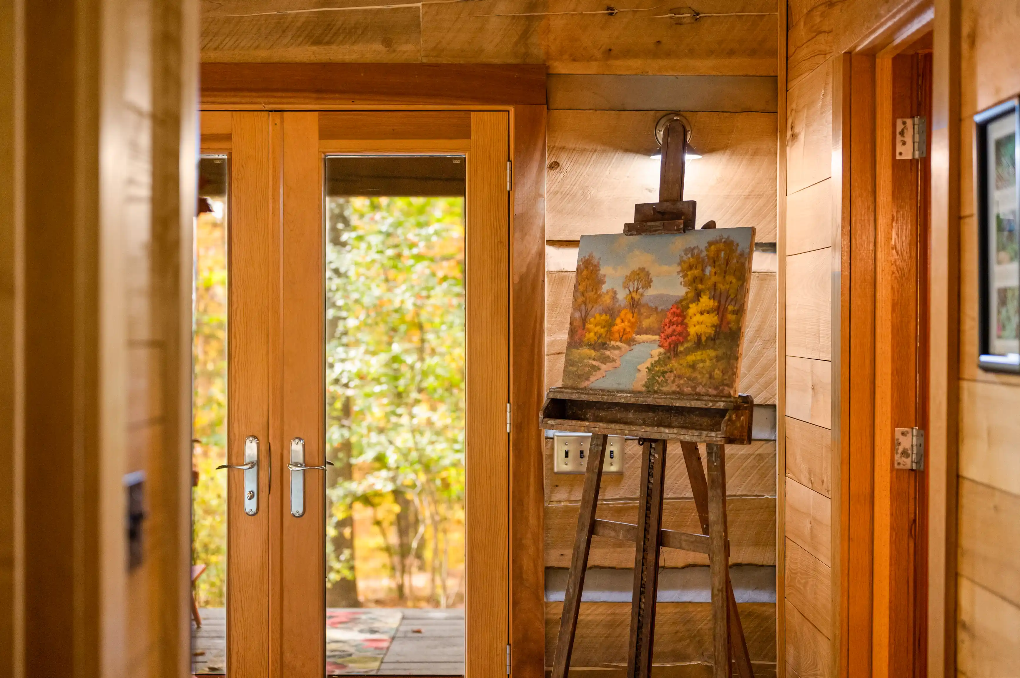 Alt text: An autumn landscape painting on an easel inside a cozy wooden room with an open door leading to the outdoors.