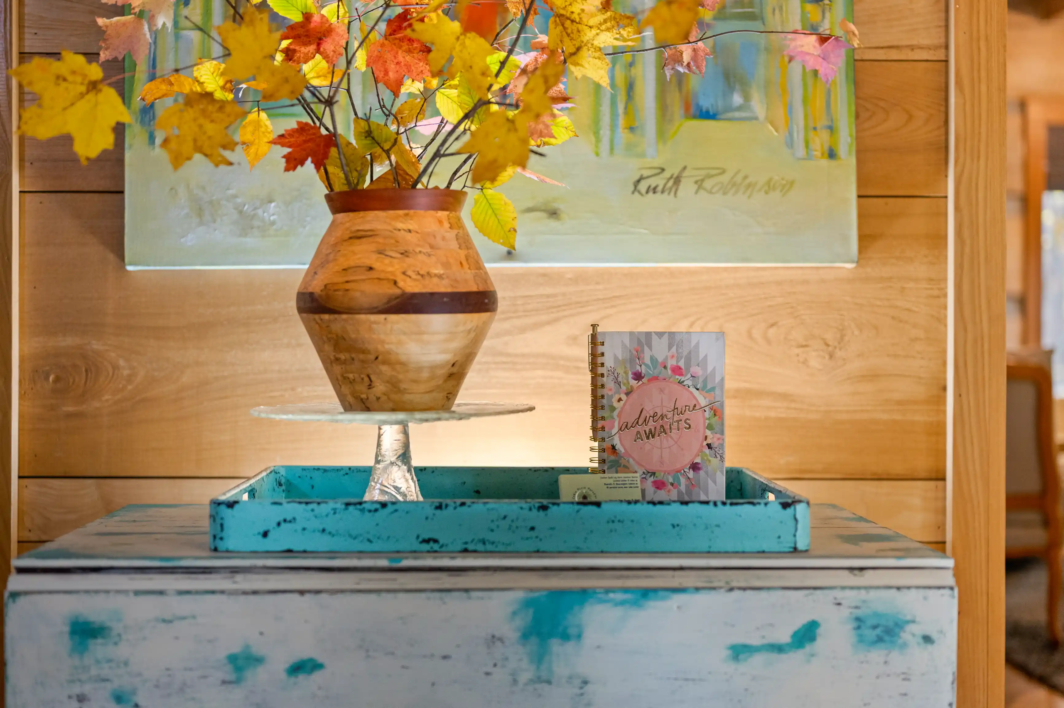 Vase with autumn leaves on a rustic blue painted wooden stand with a notebook titled 'Adventure Awaits,' beside it, in a warmly lit room with a painting in the background.