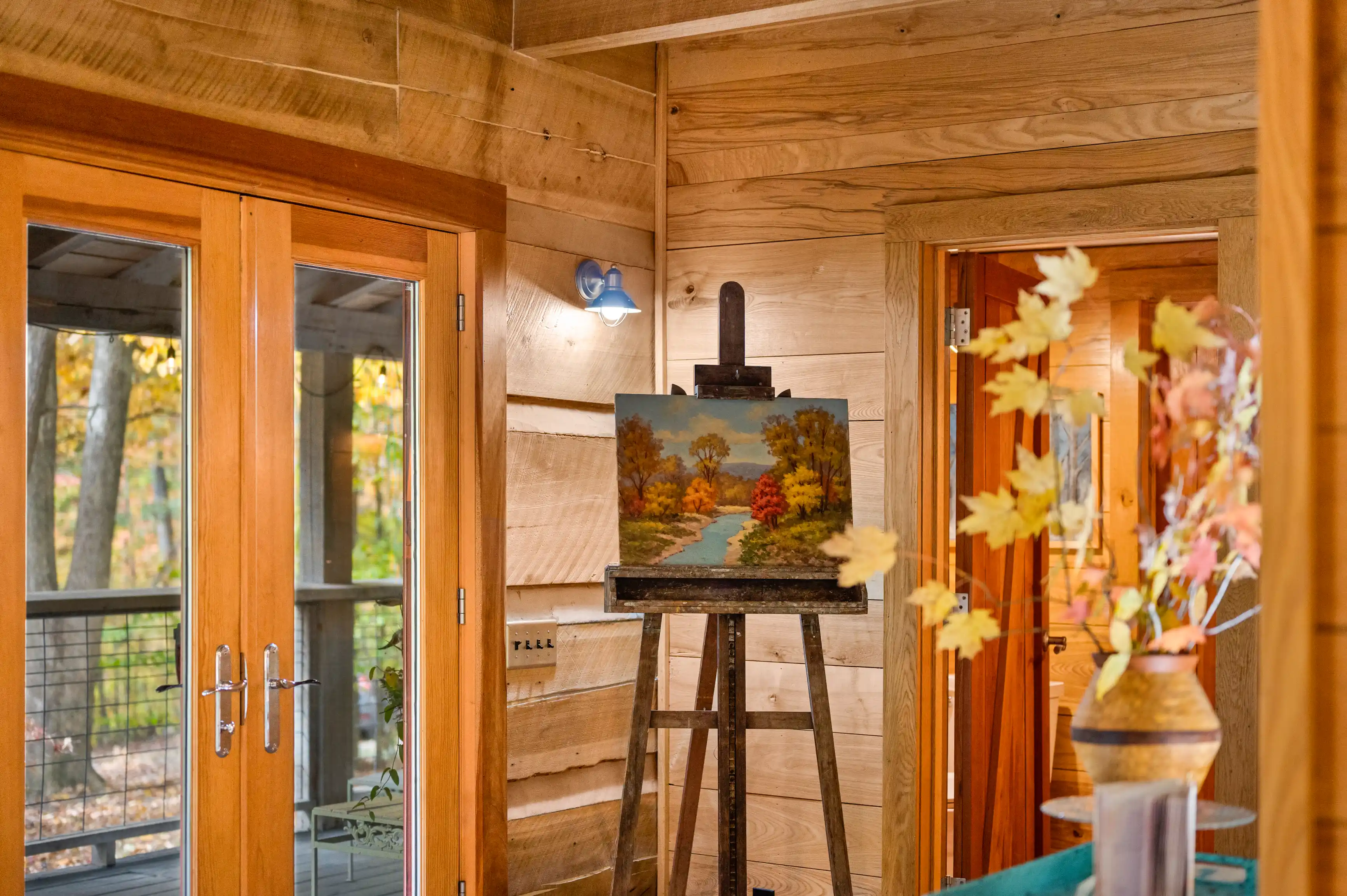 A cozy art corner in a wooden cabin with a landscape painting on an easel, a blue lamp on the wall, and glass doors opening to a deck surrounded by autumn trees.