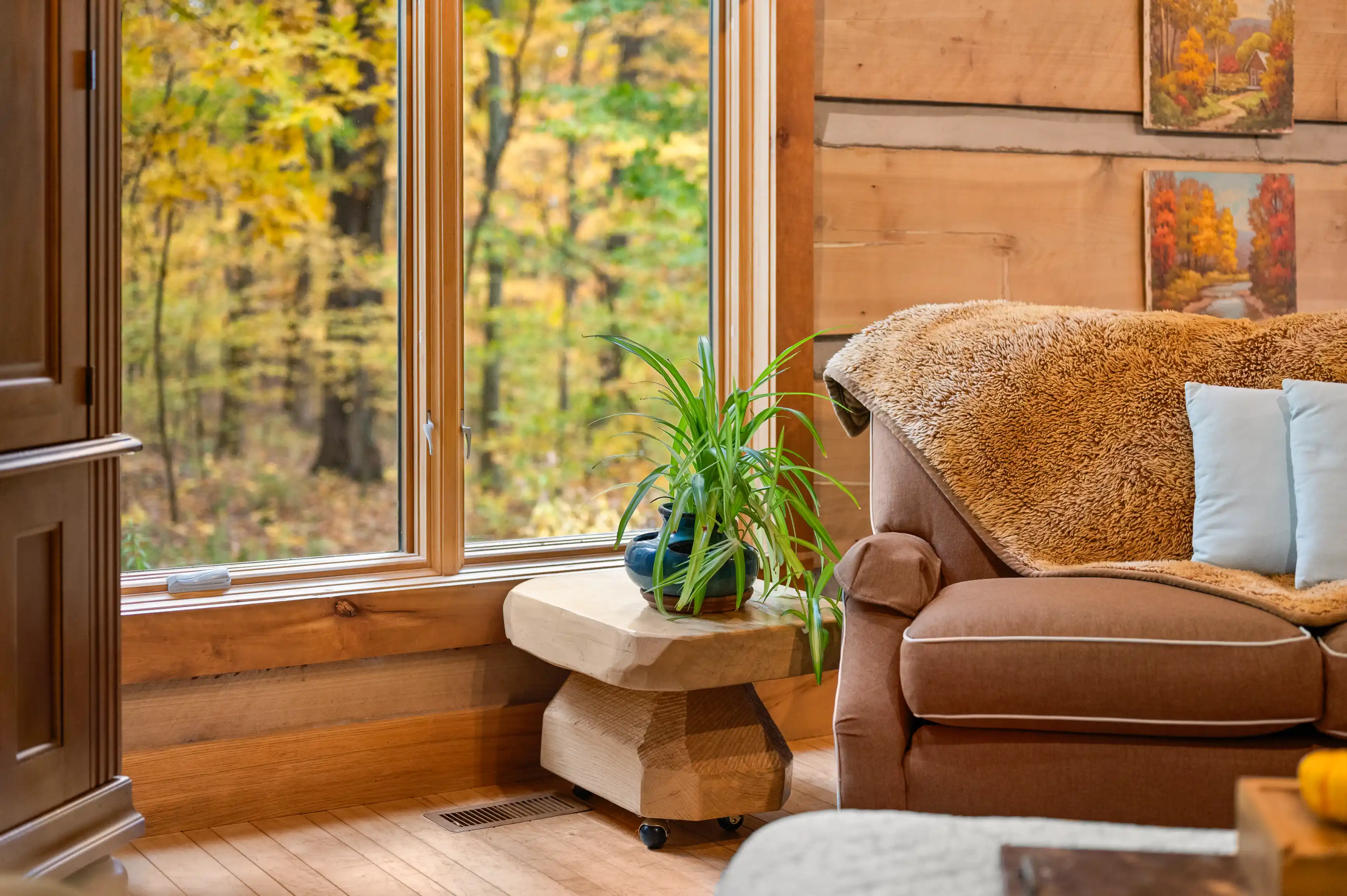 Cozy wooden cabin interior with a plush sofa, decorative pillows, a potted plant on a side table, and forest view through large windows in autumn.