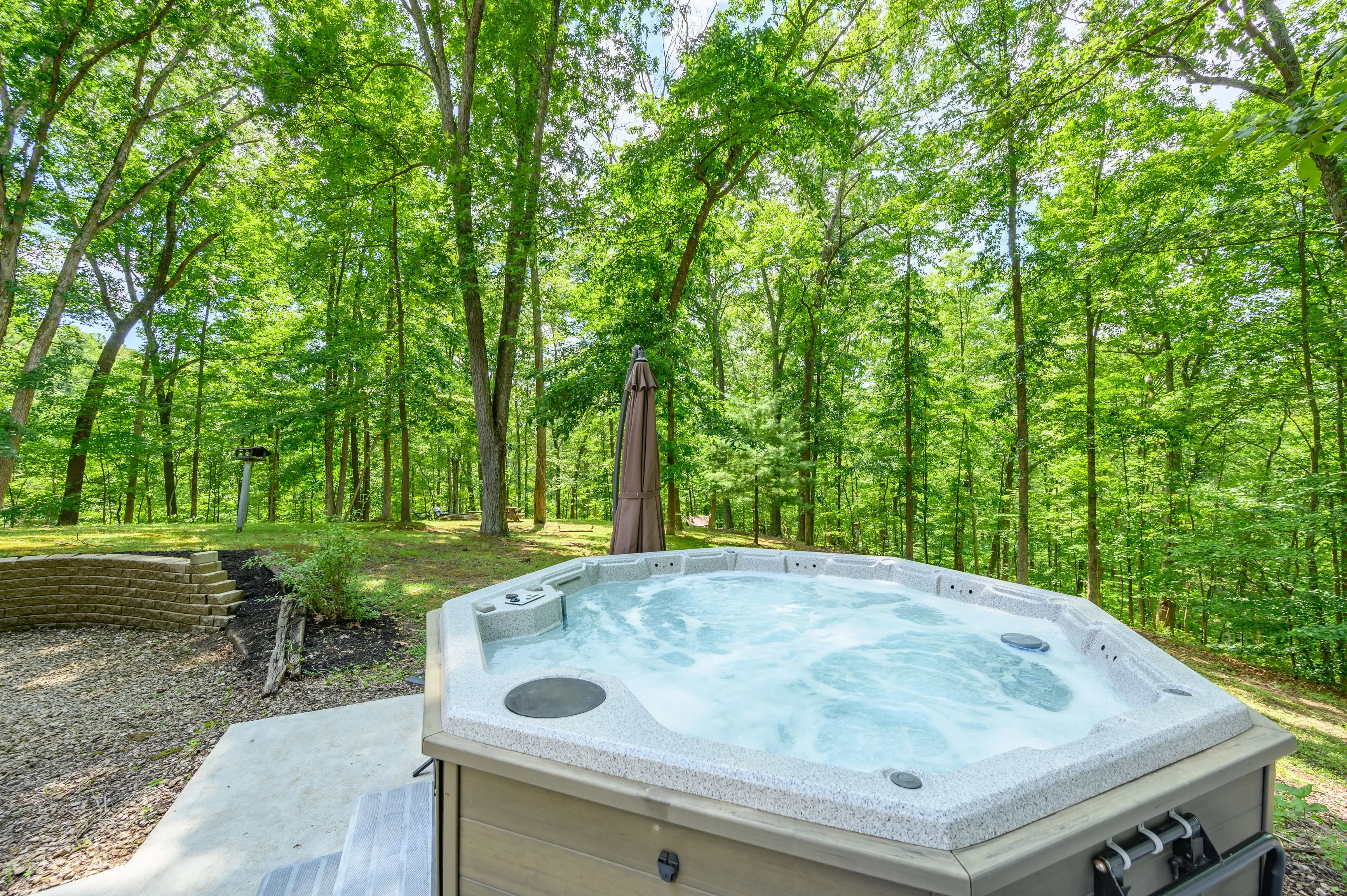 Outdoor hot tub with running water on a concrete patio surrounded by lush green woods.