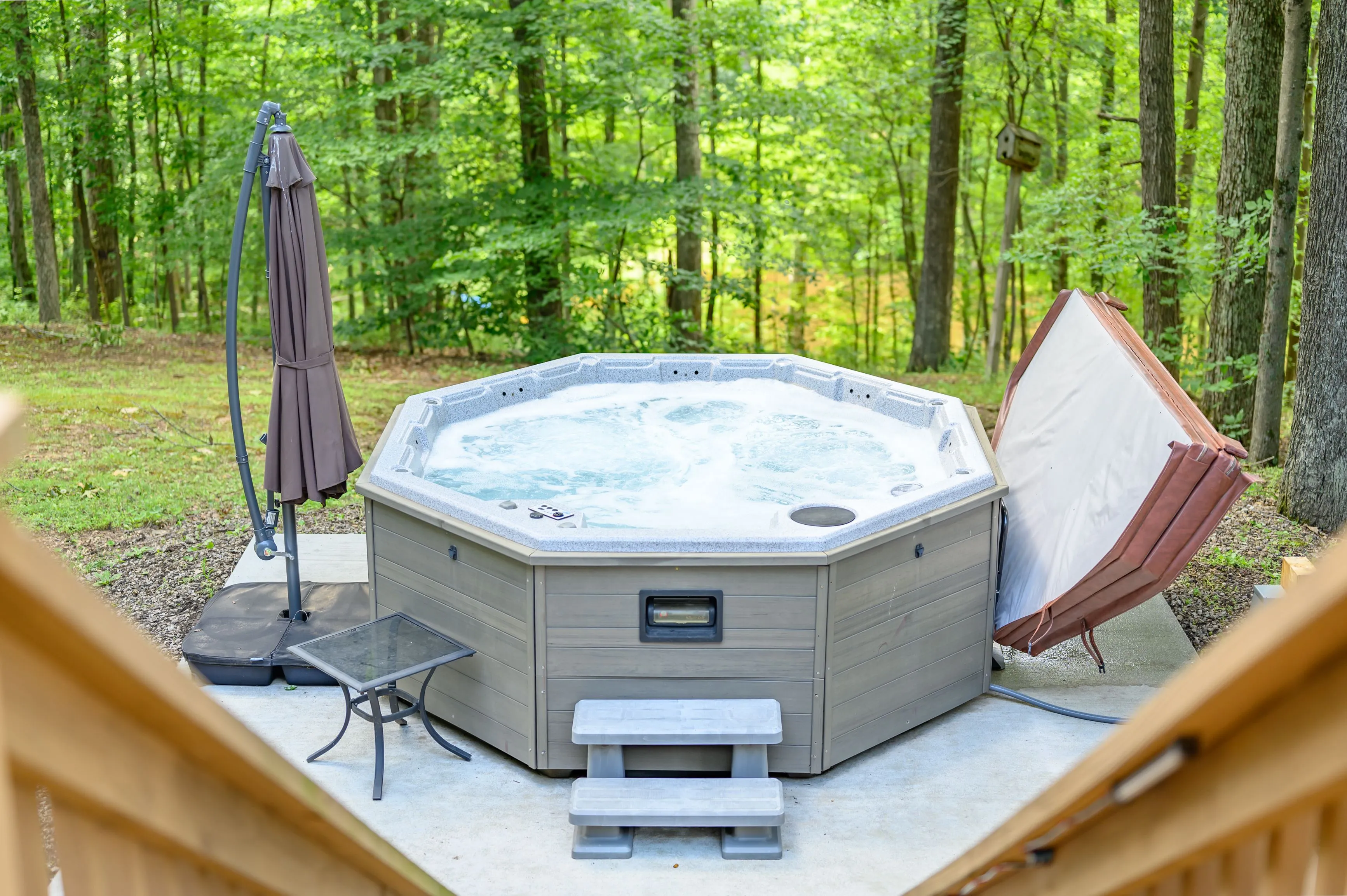 An outdoor hot tub with bubbling water on a patio surrounded by trees, with a folded lounge chair and a closed patio umbrella nearby.