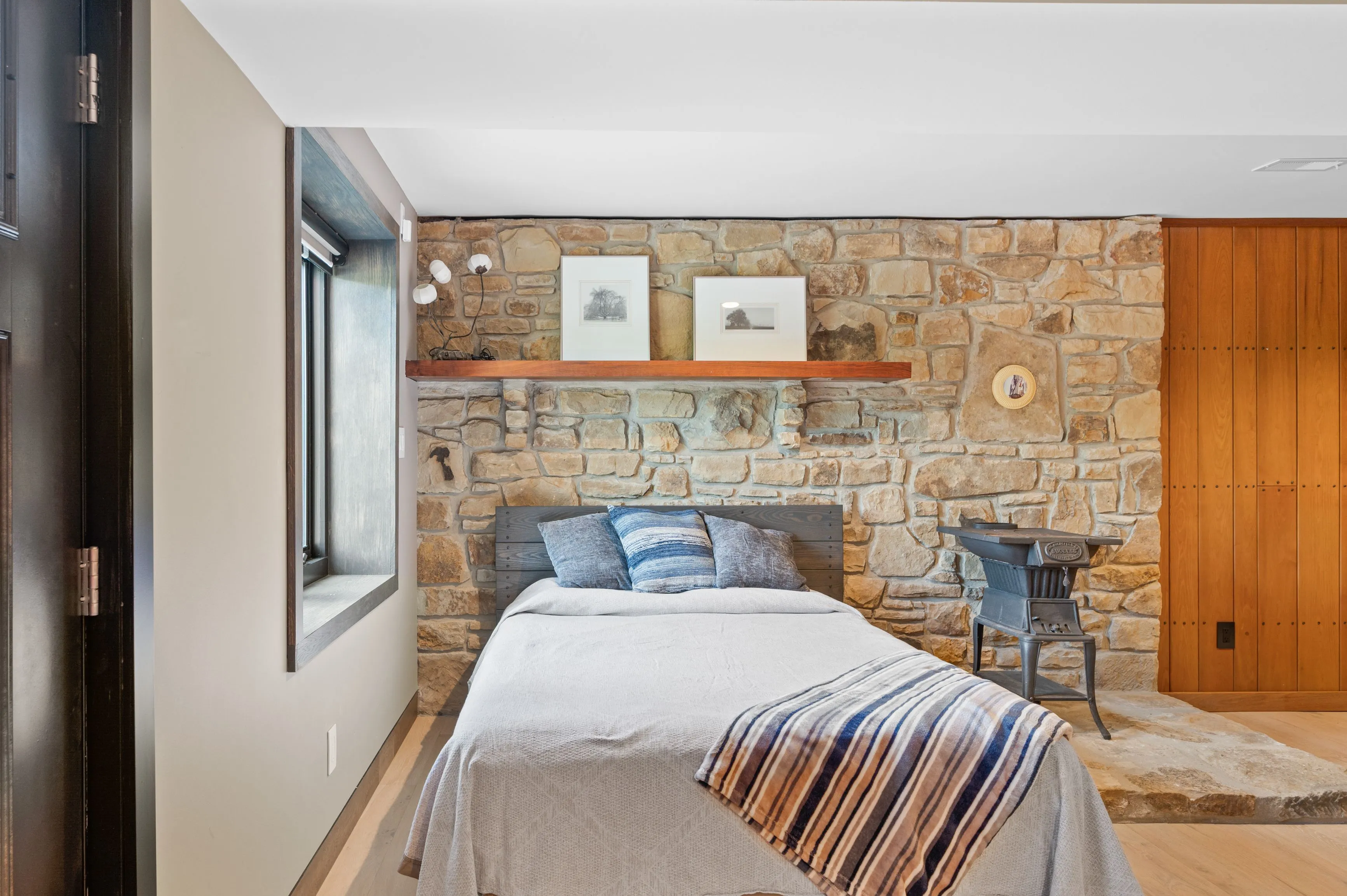 Modern bedroom interior with stone wall, wooden headboard and accents, grey bedding, and vintage wood stove.