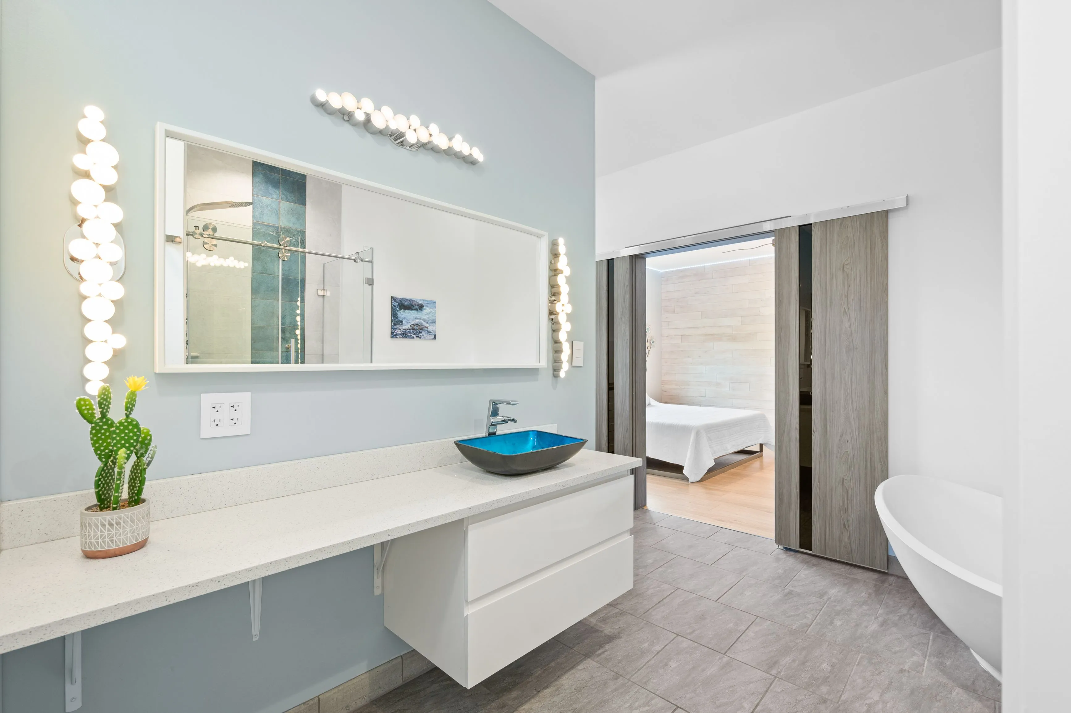 Modern bright bathroom interior with a blue vessel sink, long mirror with vanity lights, a cactus decoration, and a view into the adjoining bedroom with a freestanding bathtub.