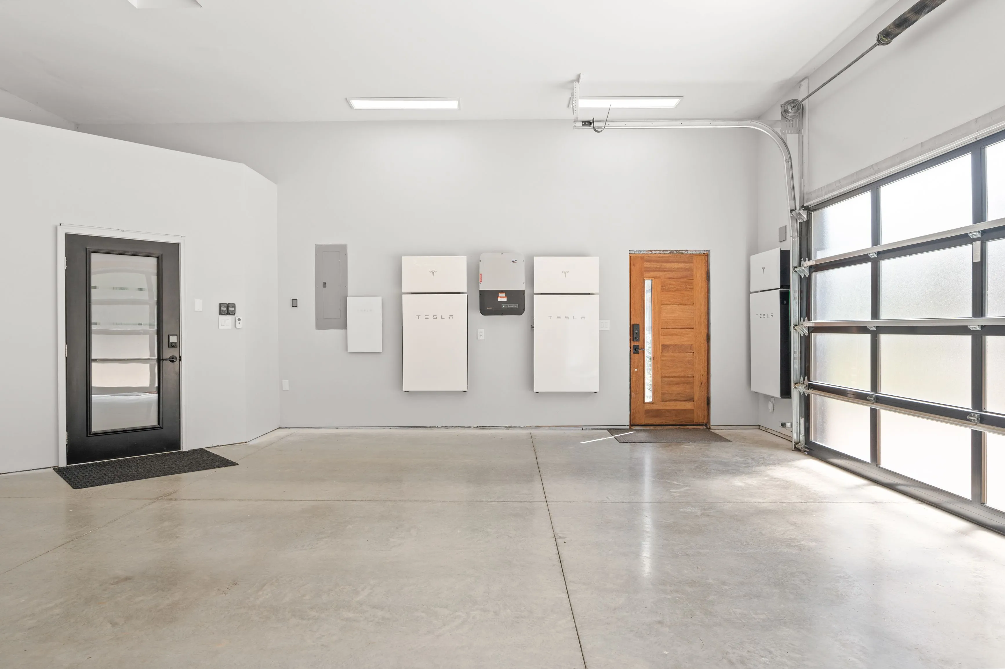 A clean and modern garage interior with a Tesla home battery setup on the wall, a wooden door, and a glass-panel garage door.