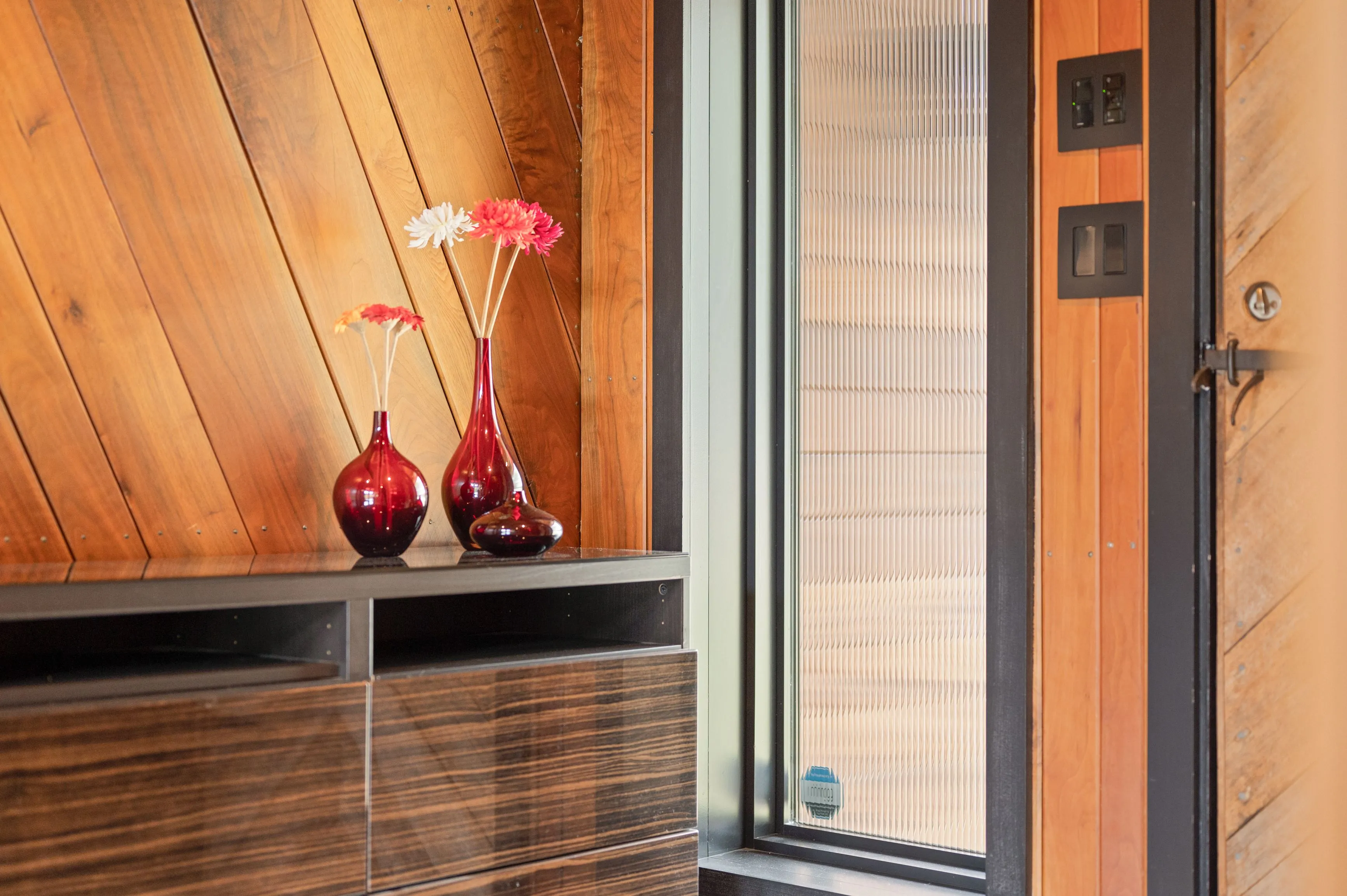 Interior corner with a wooden accent wall featuring two red vases with colorful flowers on a dark wooden credenza, next to a closed door with modern switches on the wall.