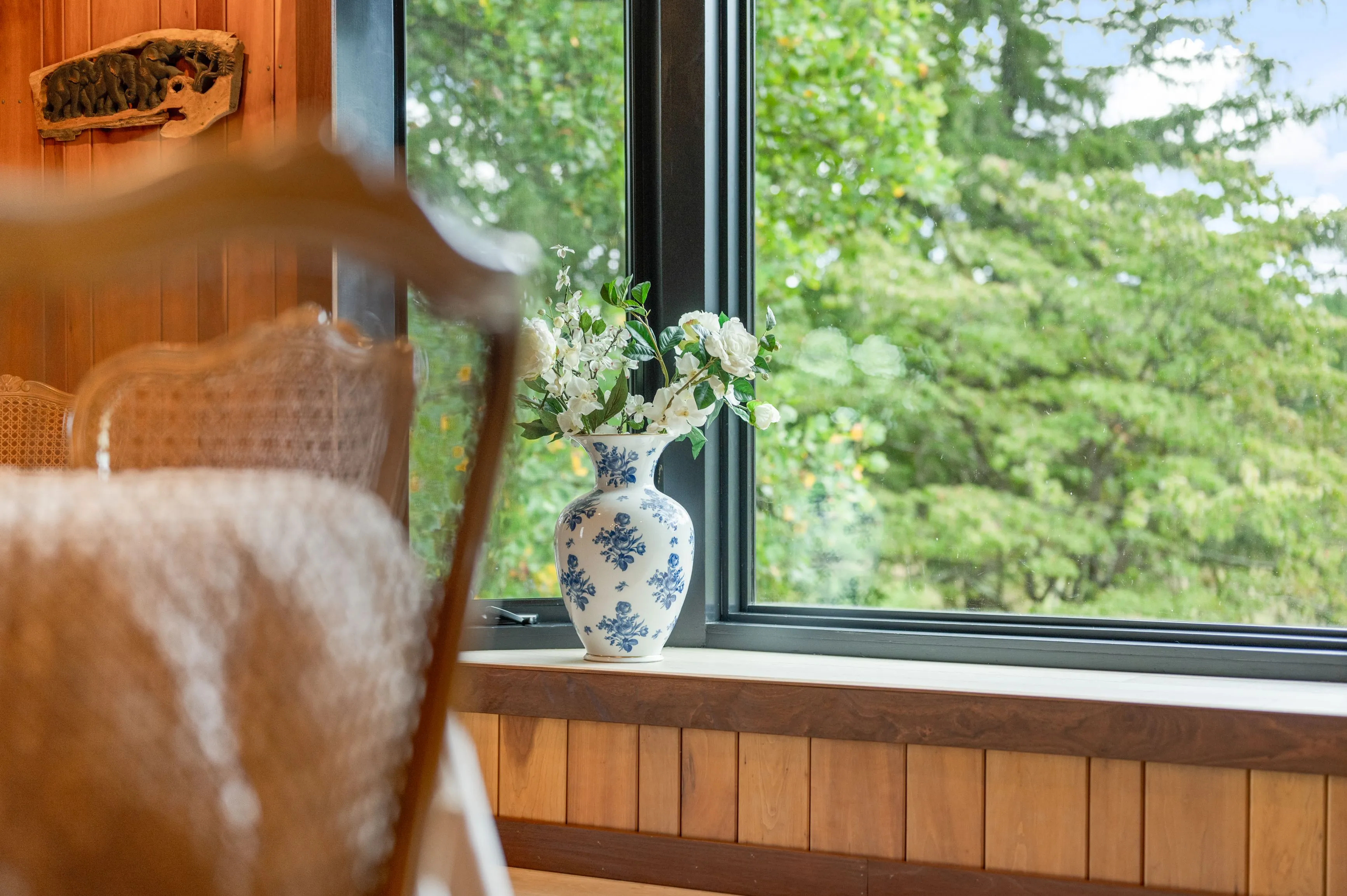 Vase with white flowers on a windowsill overlooking green trees, with partial view of a wooden chair in the foreground.
