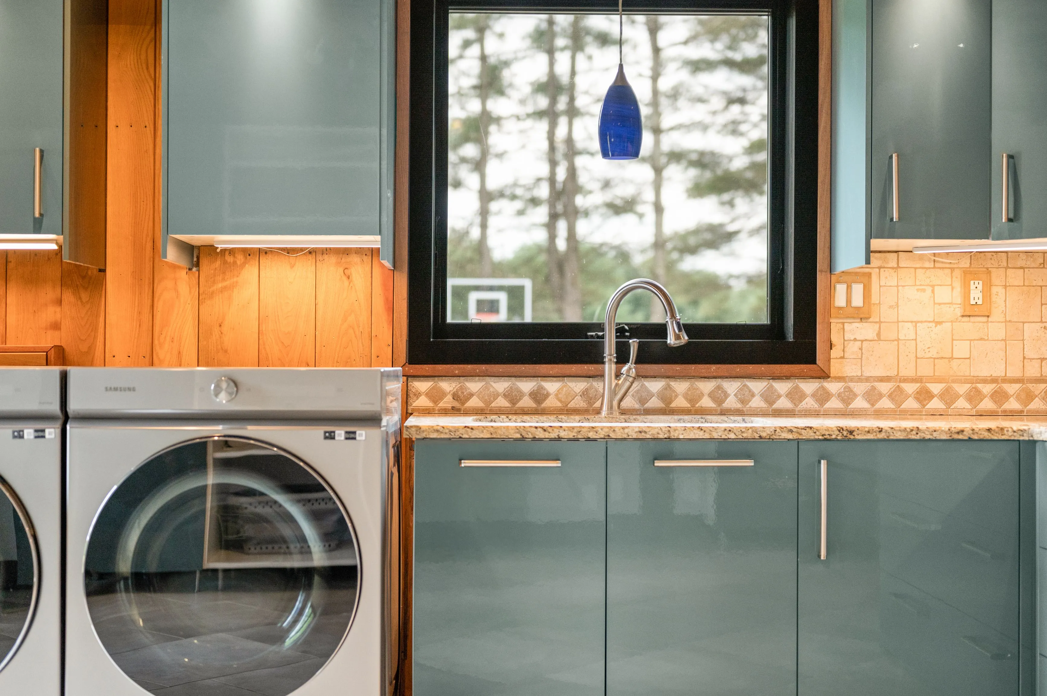 Modern laundry room featuring a front-loading washer and dryer, wooden cabinetry, and a kitchen sink with a window view.