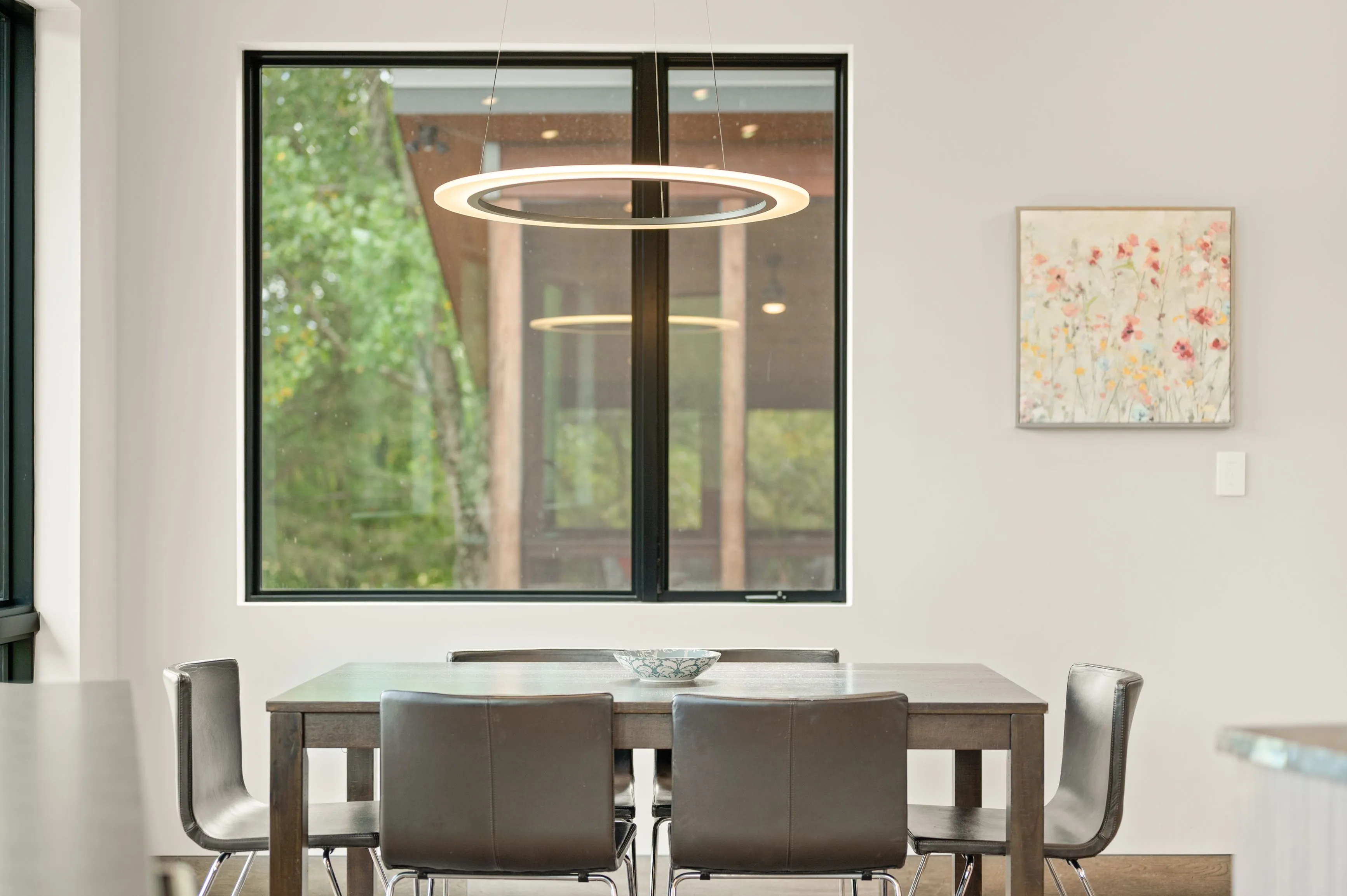 Modern dining room with a wooden table, leather chairs, hanging circular lights, and a large window overlooking trees.