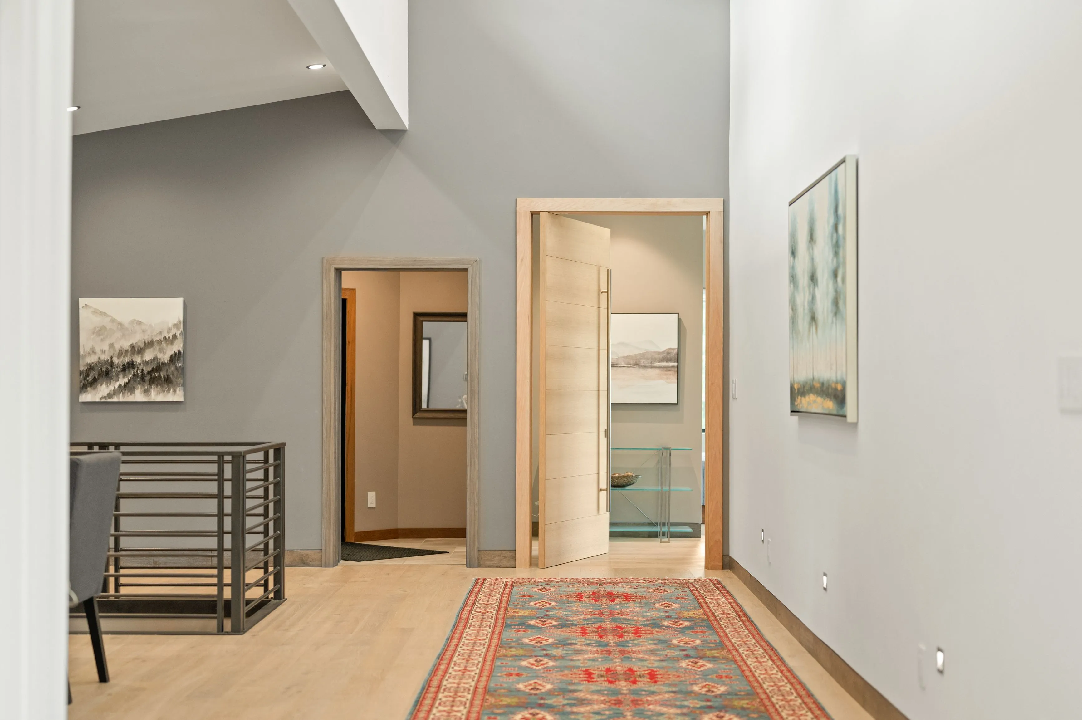 Modern home interior featuring a hallway with framed artwork on the walls, an oriental rug on the wooden floor, and open contemporary doors leading to adjacent rooms.