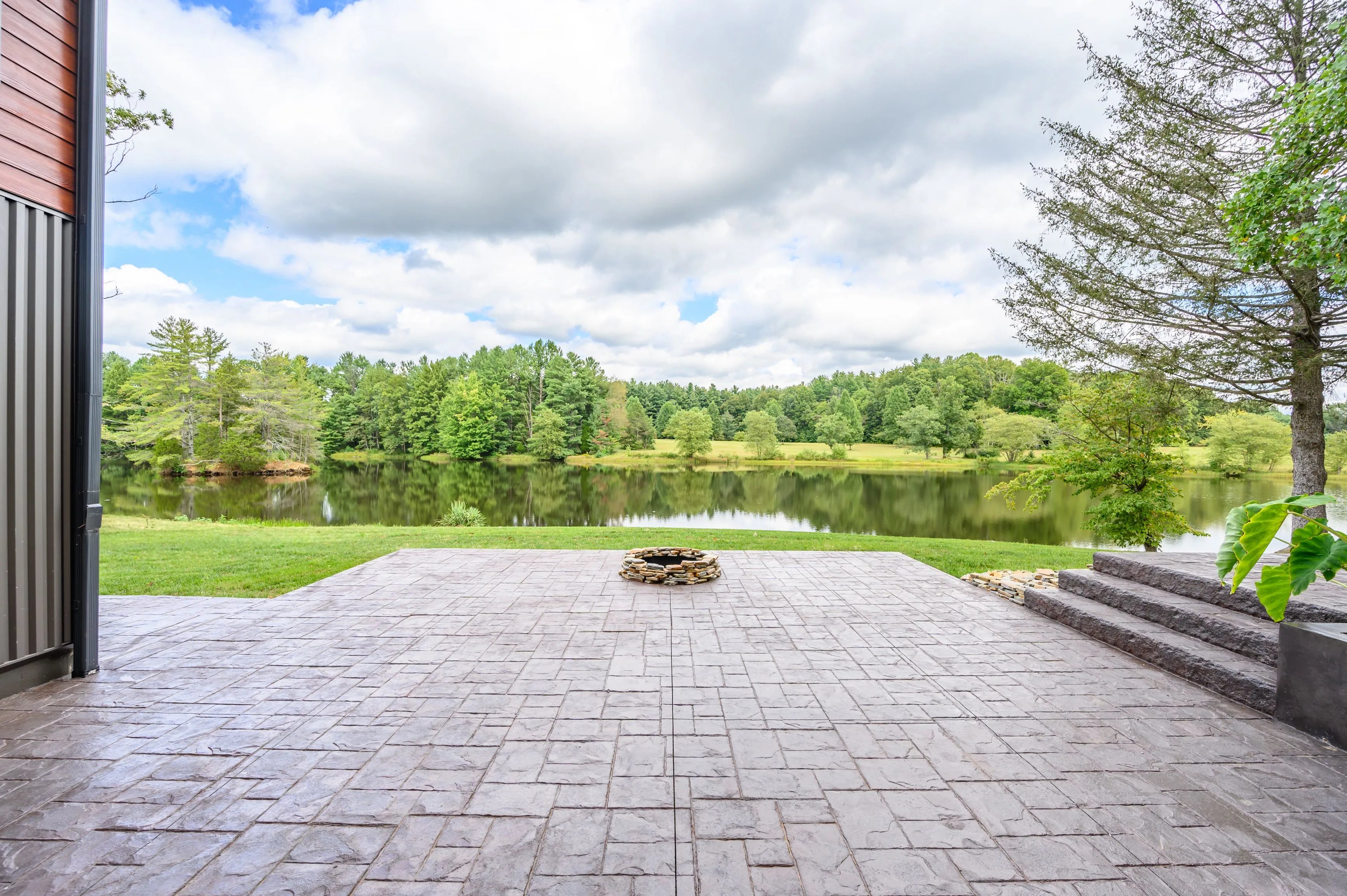 Paved patio leading to a serene lake surrounded by lush greenery and trees under a partly cloudy sky.