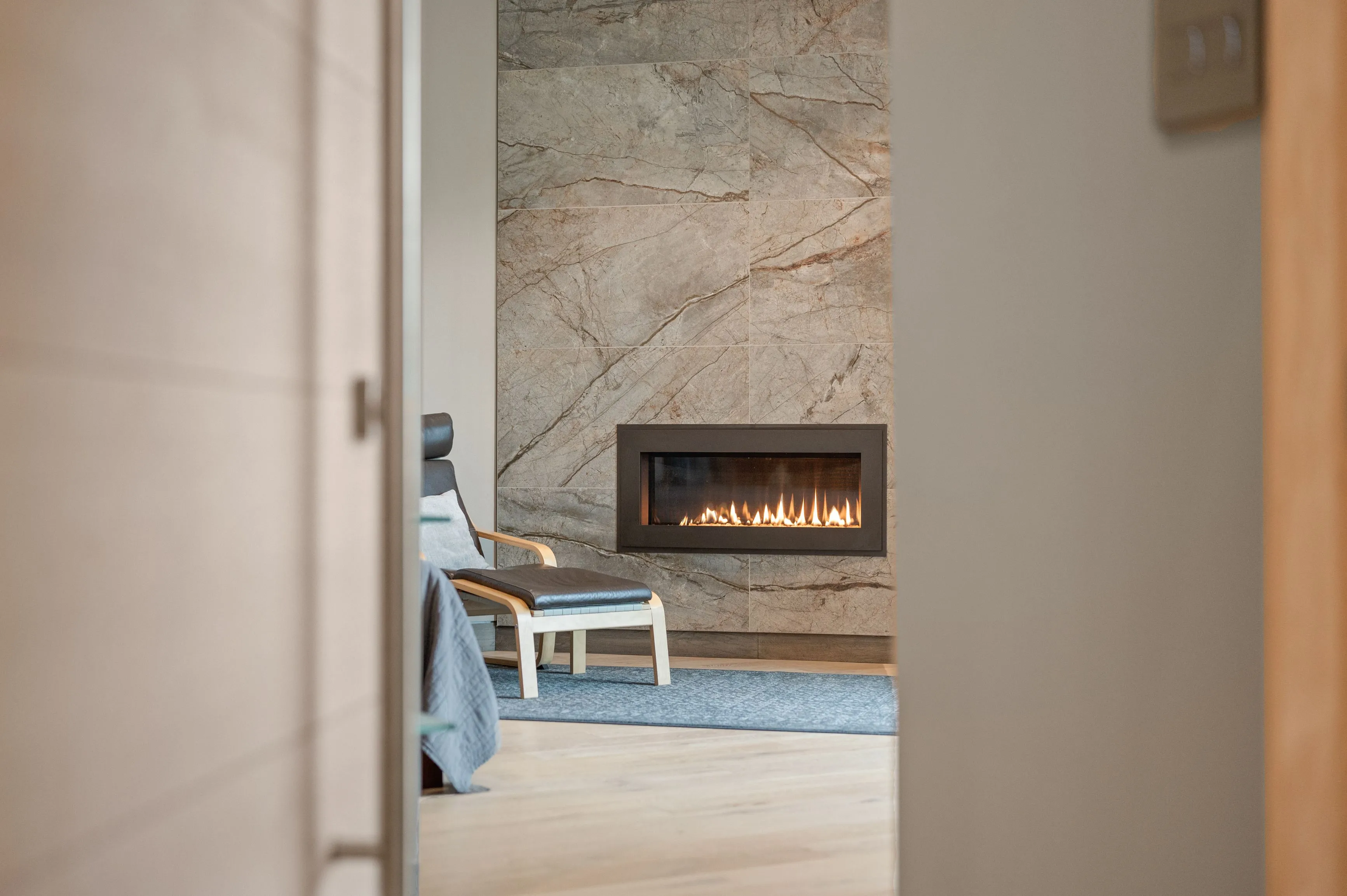 View from a partially open door towards a modern living room with a comfortable chair and lit gas fireplace set in a stone wall.