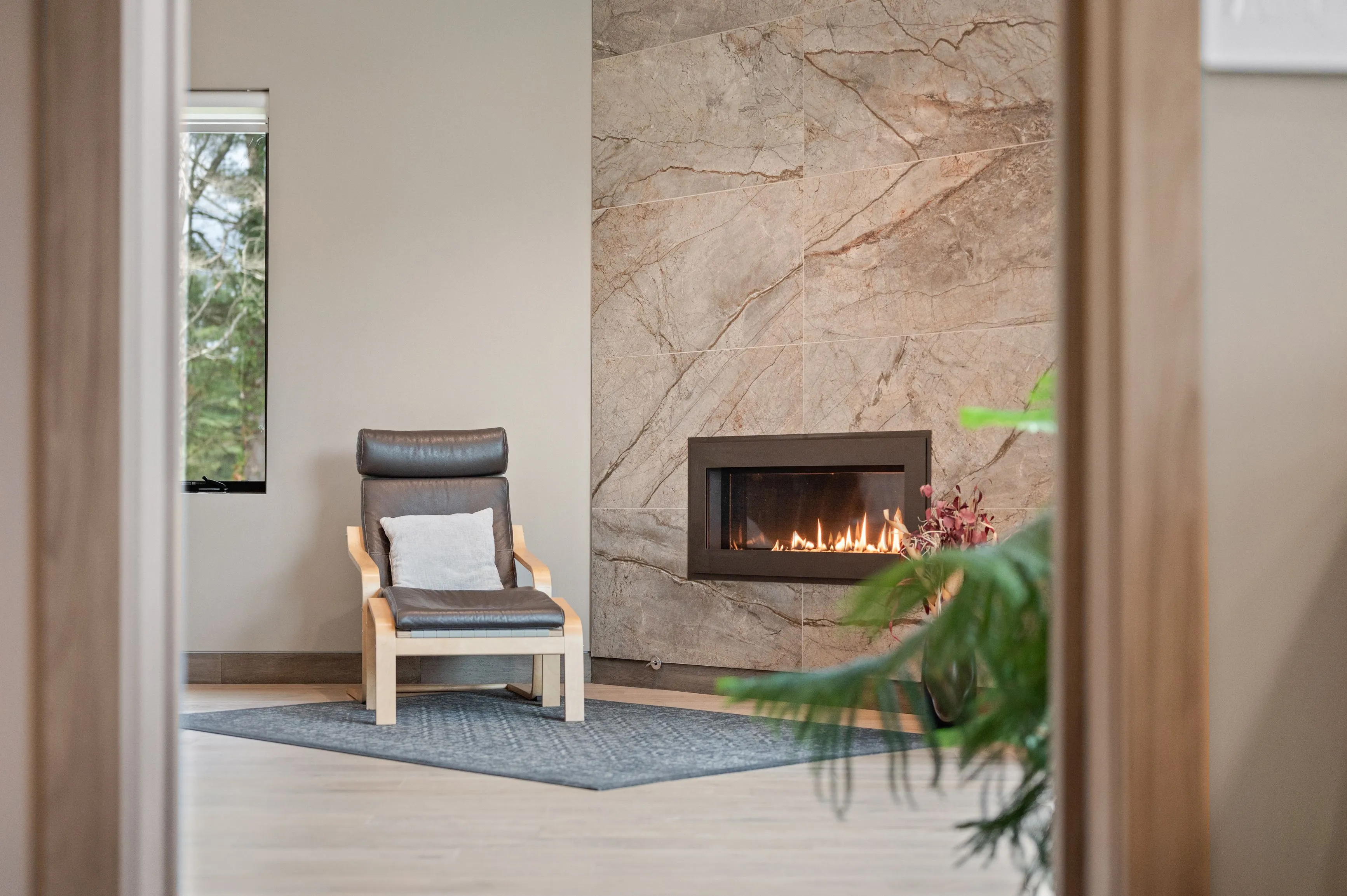 Modern cozy living room with a leather armchair, a lit fireplace surrounded by natural stone tiles, and a view of greenery outside the window.