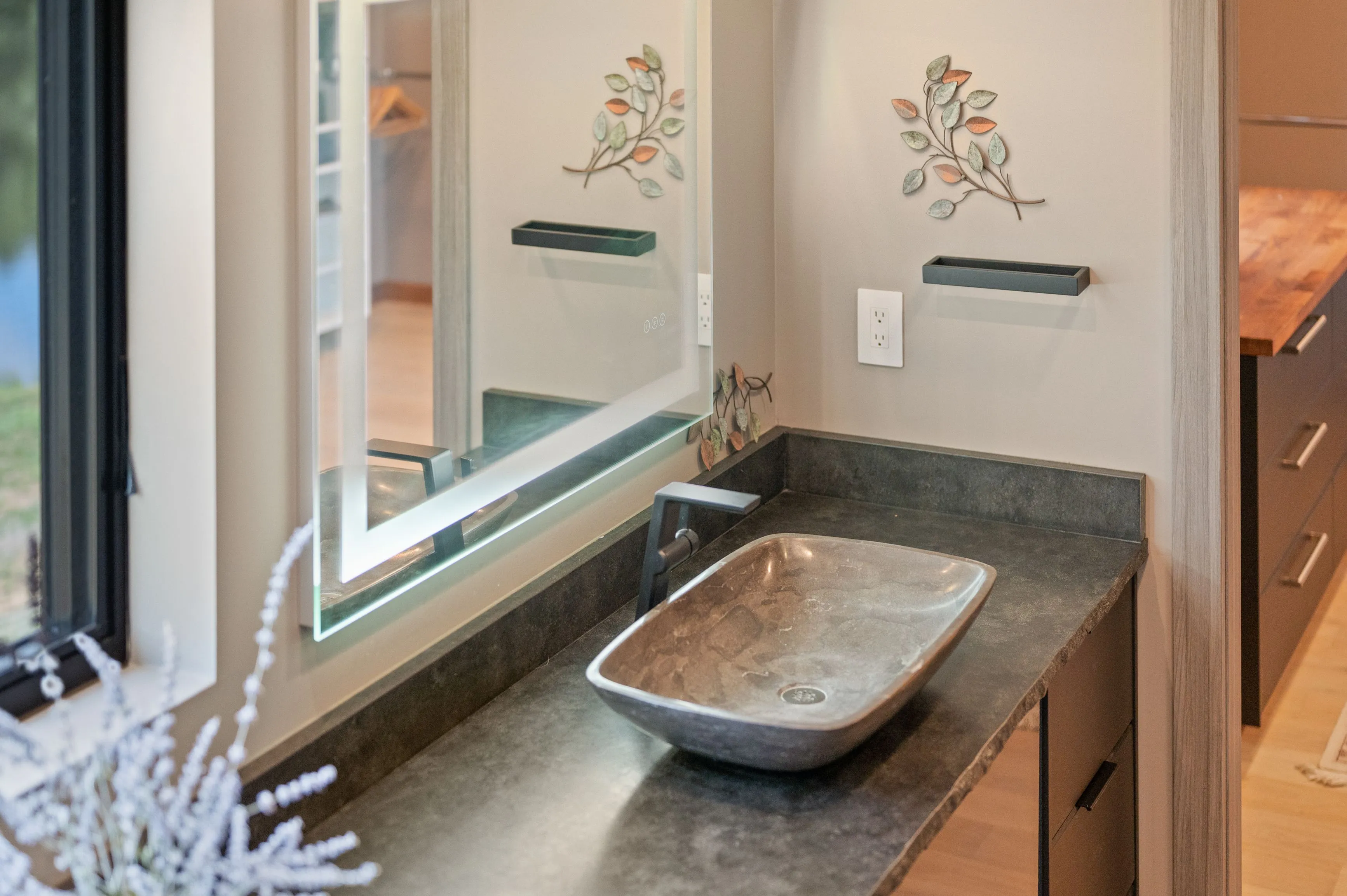 Modern bathroom with a stone countertop and vessel sink, decorative mirror, and wall-mounted shelf, situated beside a window with a view of trees.