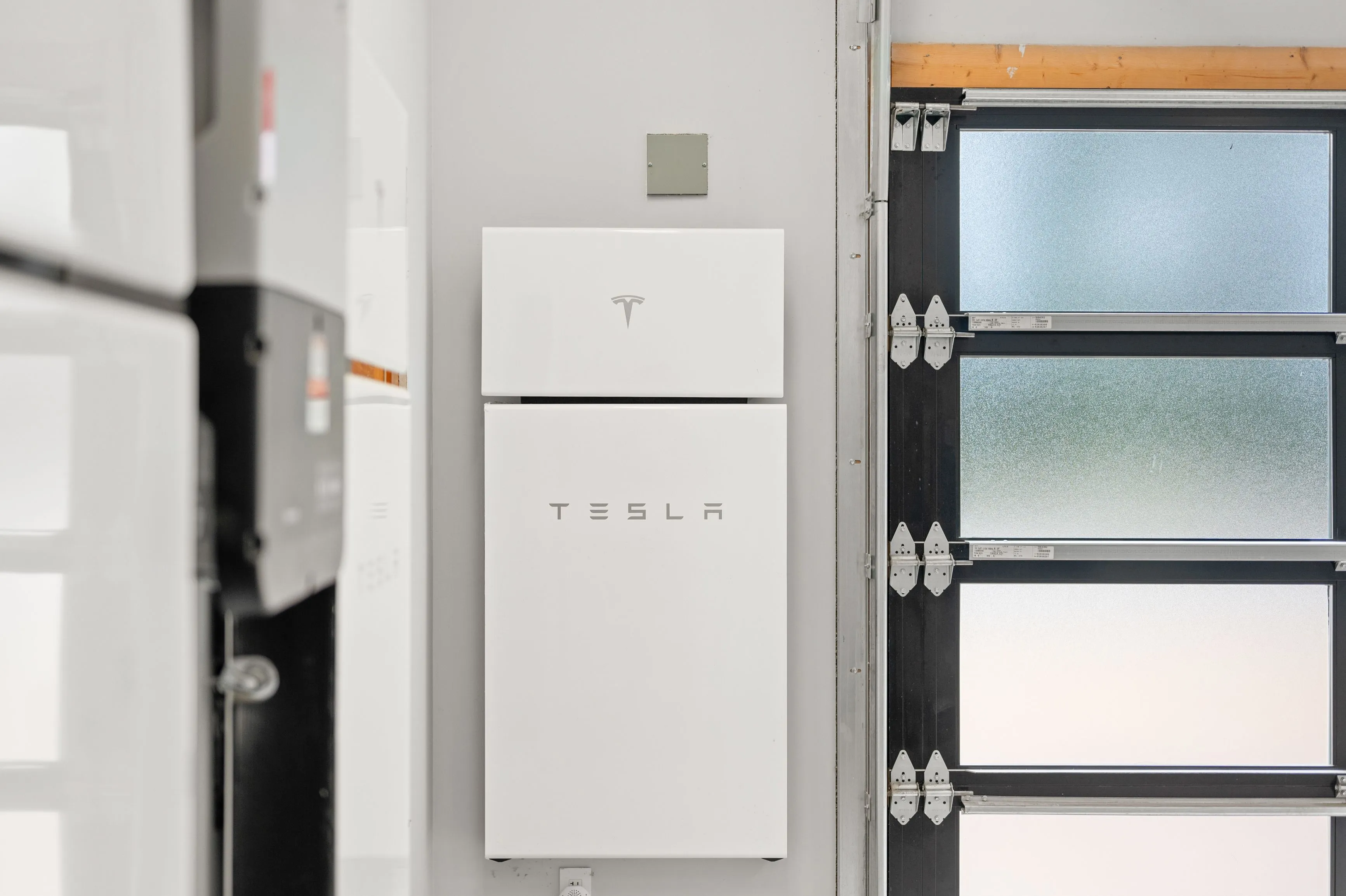 Tesla Powerwall battery storage unit mounted on a wall next to frosted glass windows in a modern home's utility area.