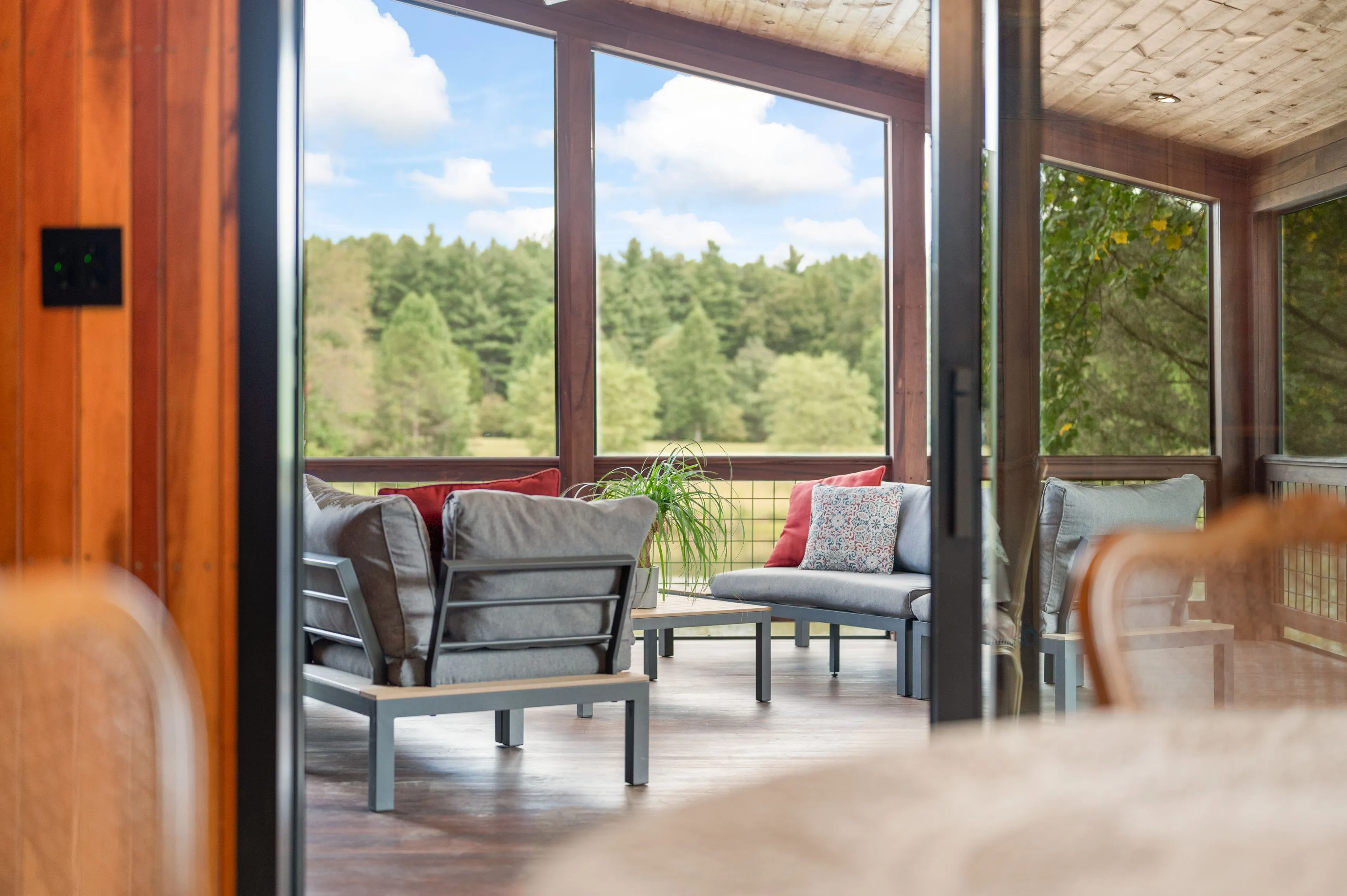 View from an open door of a cozy sunroom with modern furniture and large windows overlooking a lush forest.
