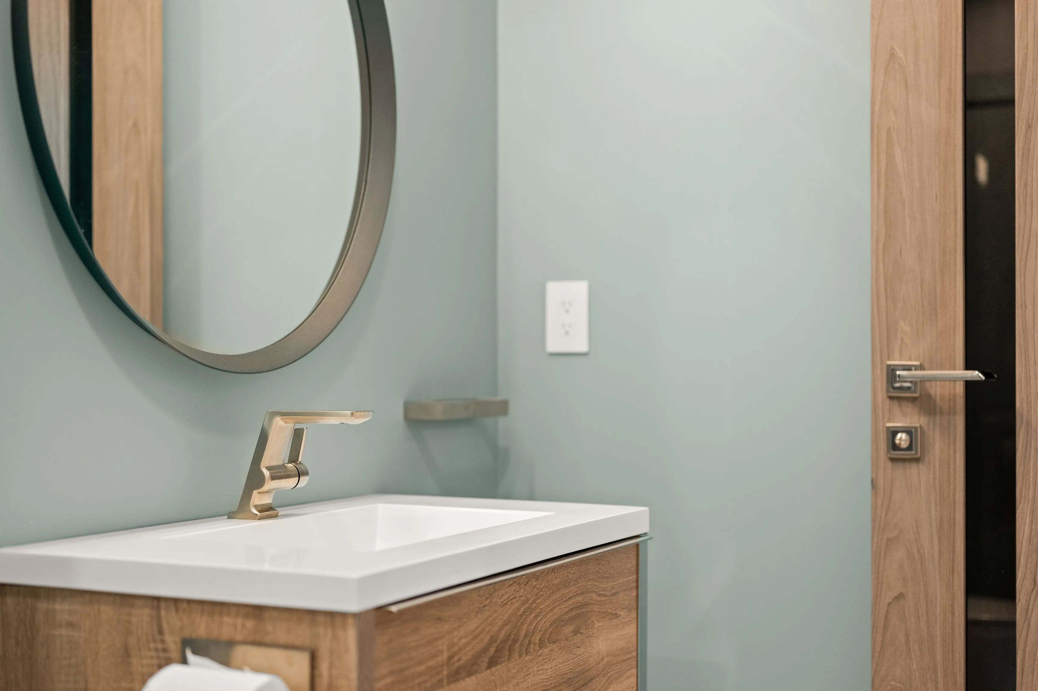 Modern bathroom with a square sink, bronze faucet, round mirror, wooden cabinet, and teal walls.