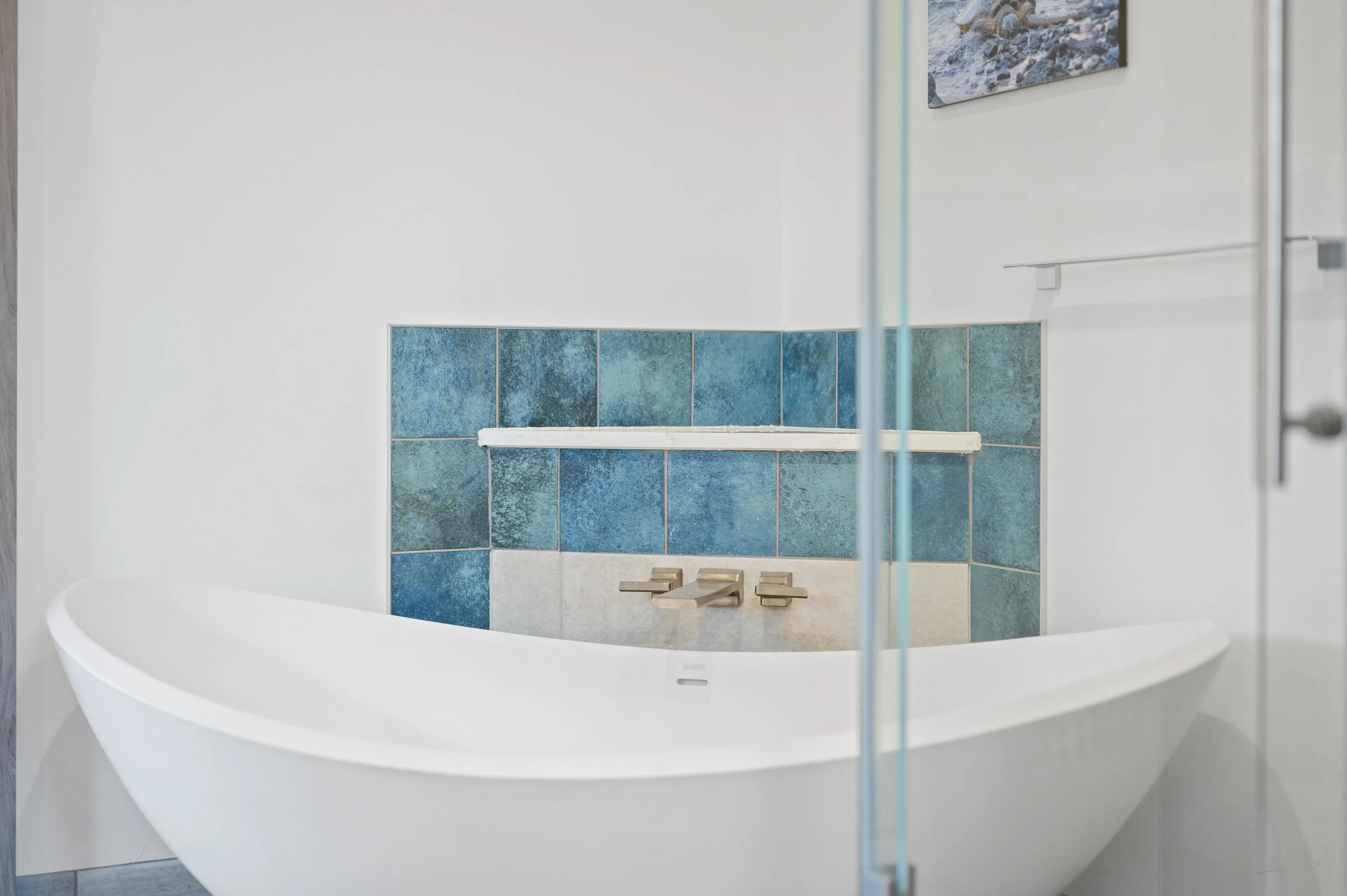 Modern bathroom interior with a white freestanding bathtub, blue and beige tiled wall, and a glass panel with a framed picture hanging on the wall.