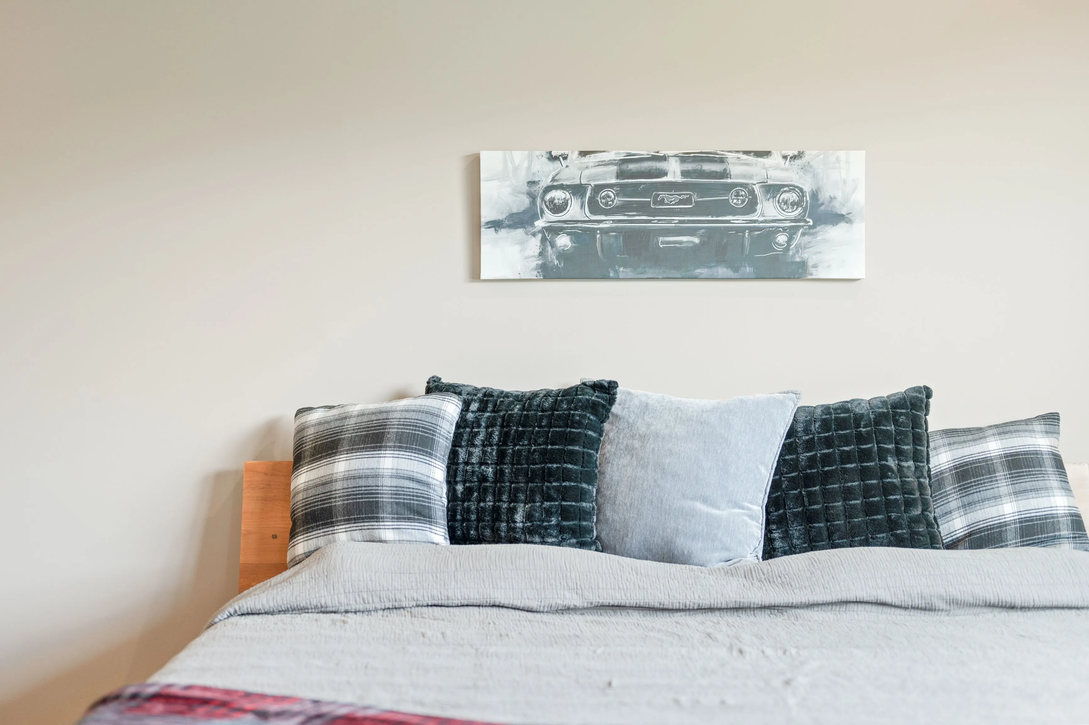 A neatly made bed with plaid and solid-colored pillows against a wooden headboard, with a grayscale artwork of a classic car hanging above on a light-colored wall.