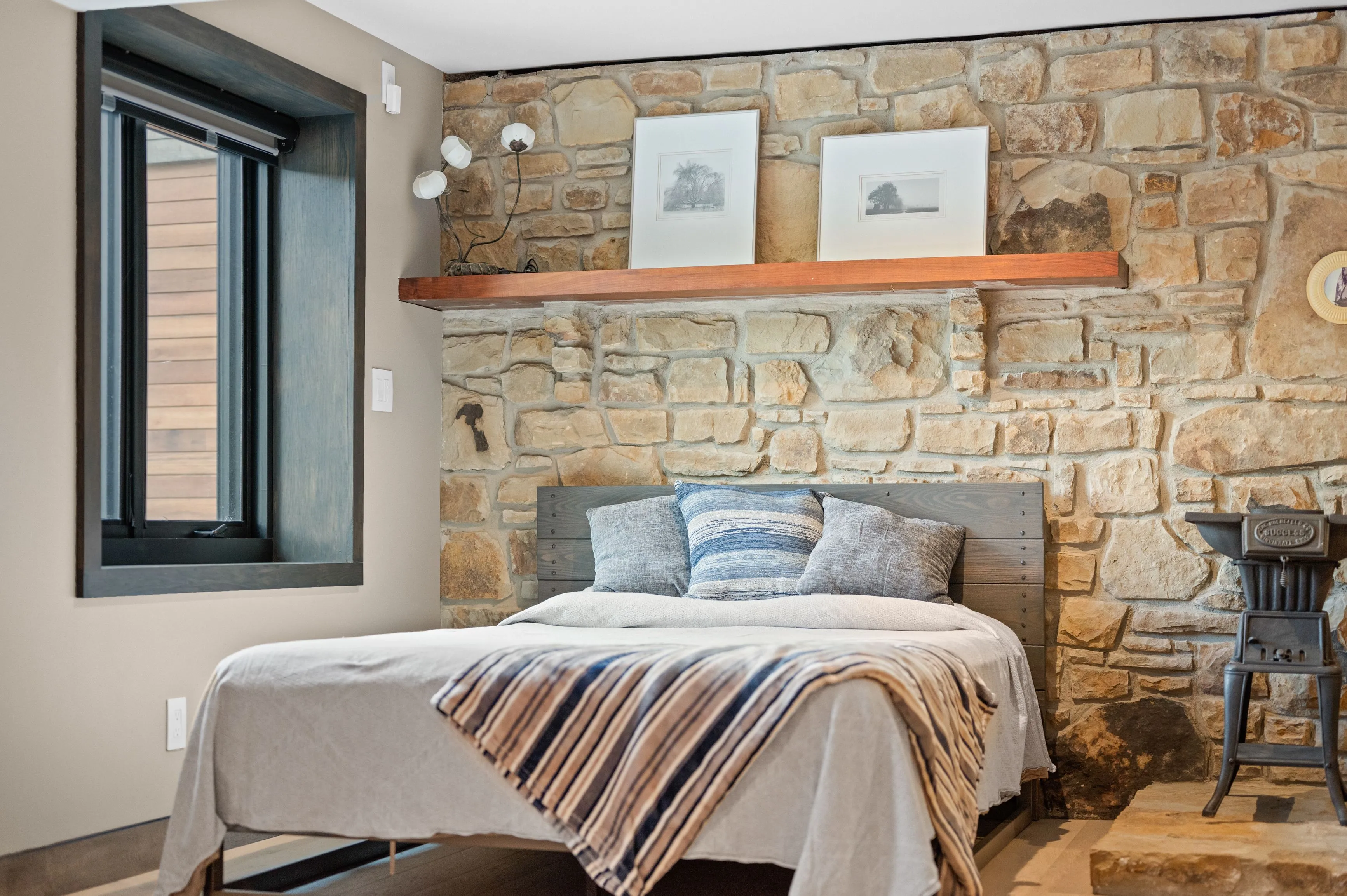 Cozy bedroom interior with stone wall, wooden shelf with framed pictures, bed with pillows and striped throw, and vintage stove on the side.