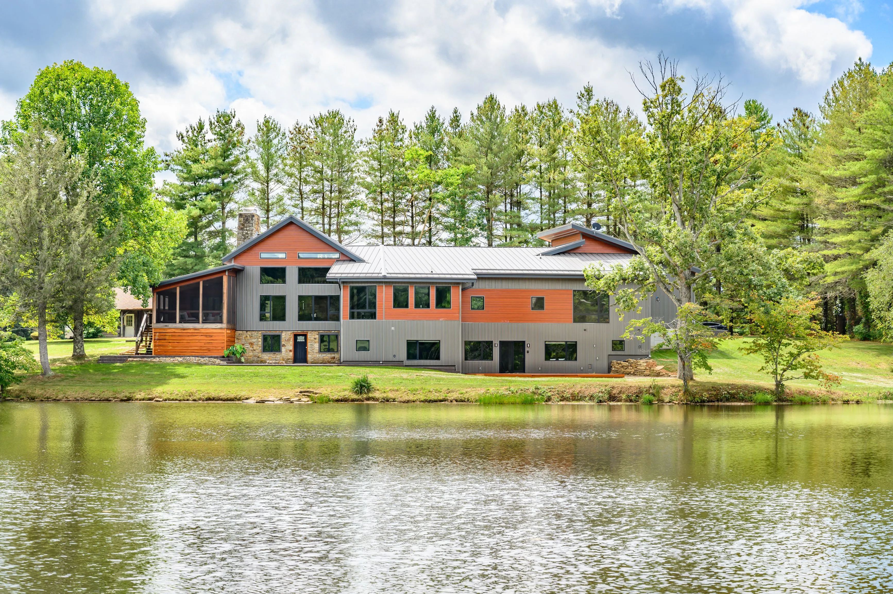 Modern multi-story house with large windows and a mix of wood and metal siding, situated by a pond surrounded by lush greenery.