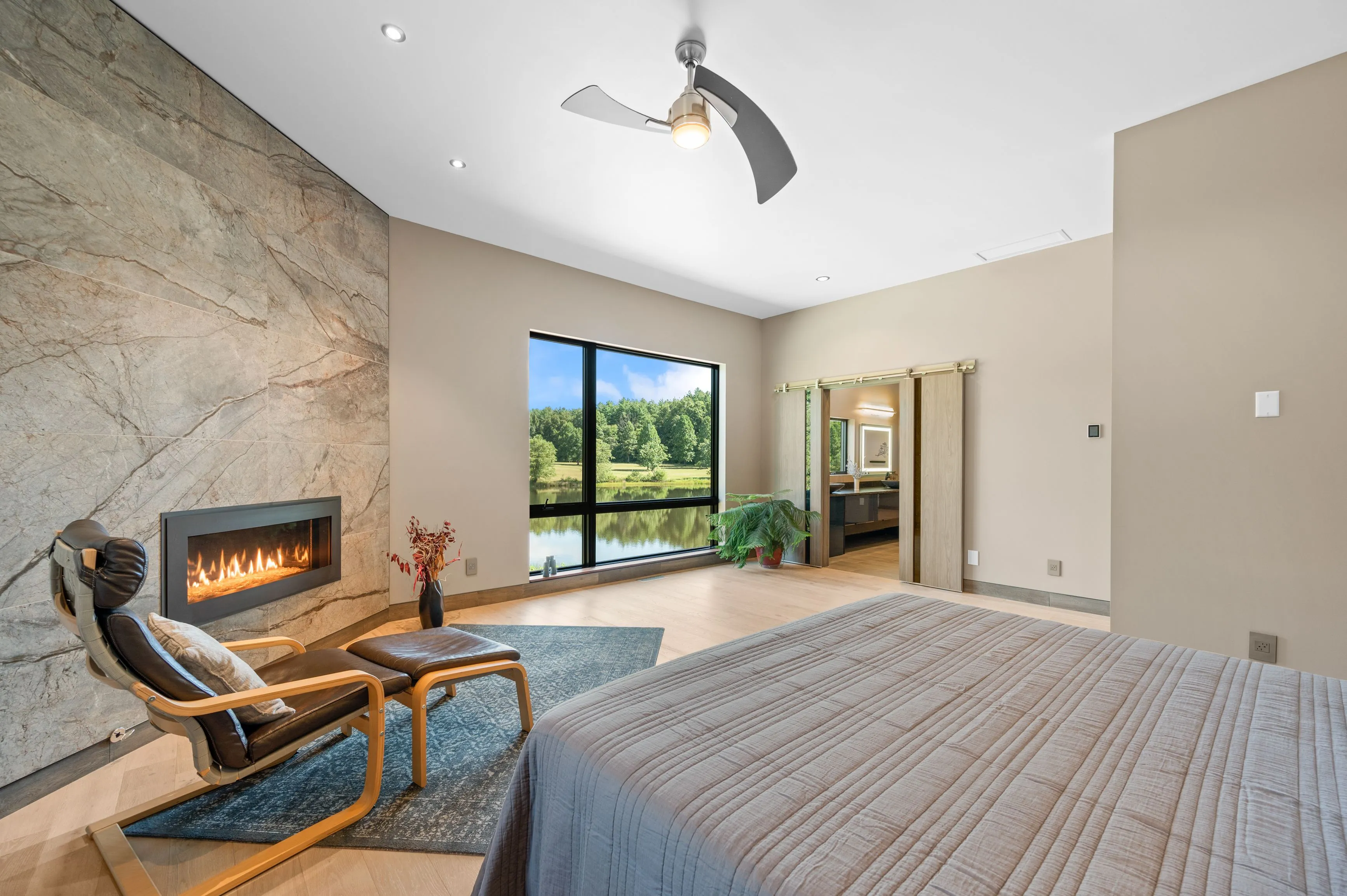 Modern bedroom interior with stone fireplace, hardwood flooring, and large window overlooking a serene lake view.